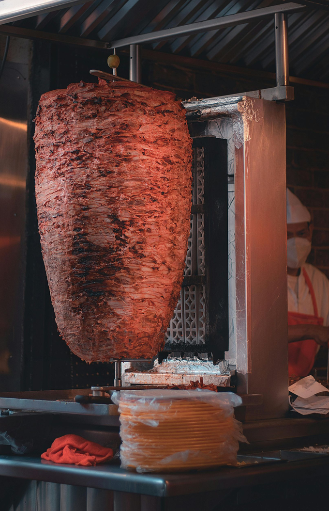 Gran cono giratorio de carne sazonada (al pastor) siendo raspada para tacos. Cocinero con bata blanca al fondo.