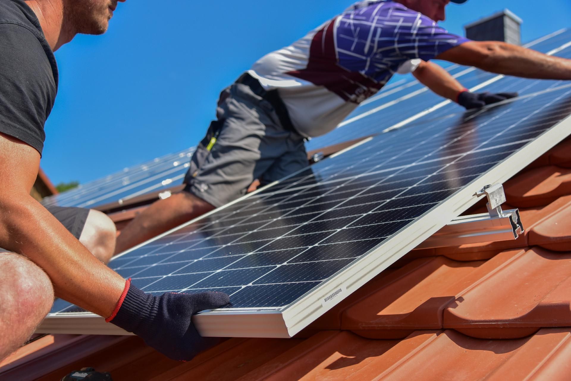 Zwei Arbeiter installieren Solarpaneele auf einem roten Ziegeldach unter strahlend blauem Himmel.