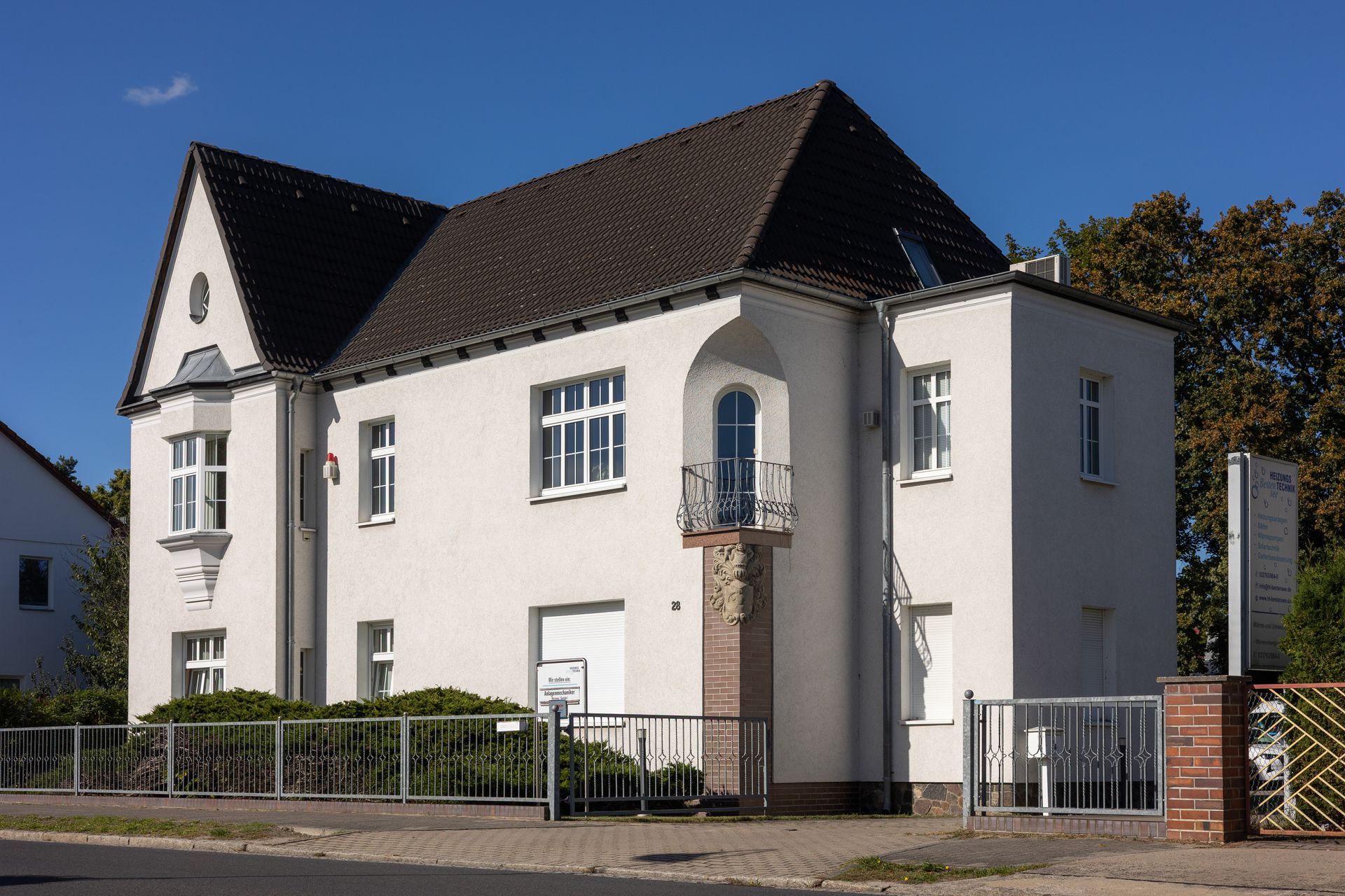 Zweistöckiges weißes Gebäude mit dunklem Dach. Ein kleiner Balkon und ein Eisenzaun sind vorhanden.