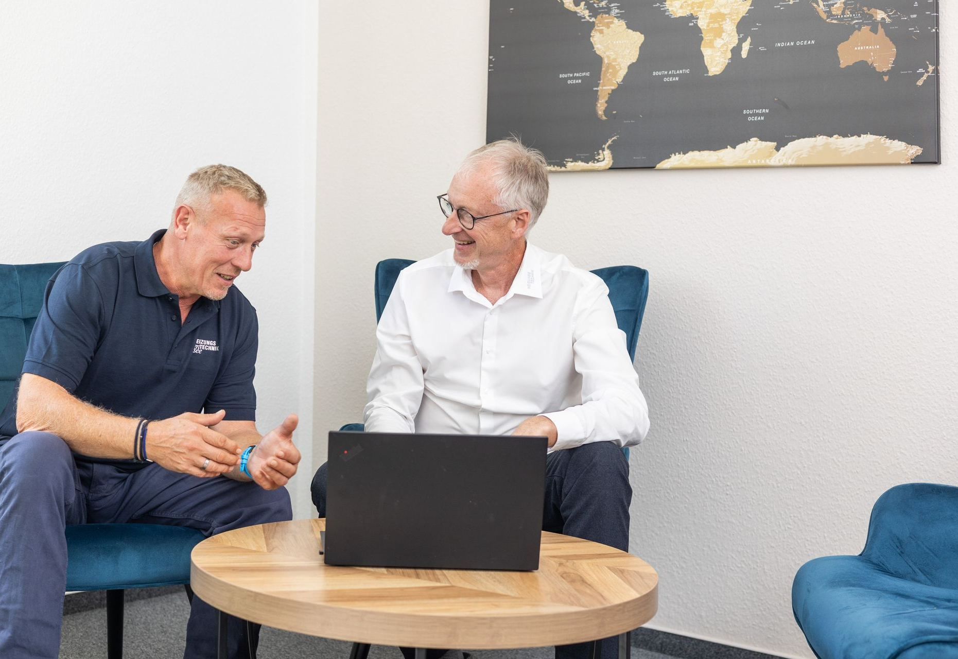 Zwei Männer sitzen in einem Geschäftsumfeld vor einem Laptop. Der eine trägt ein Poloshirt, der andere ein weißes Hemd.