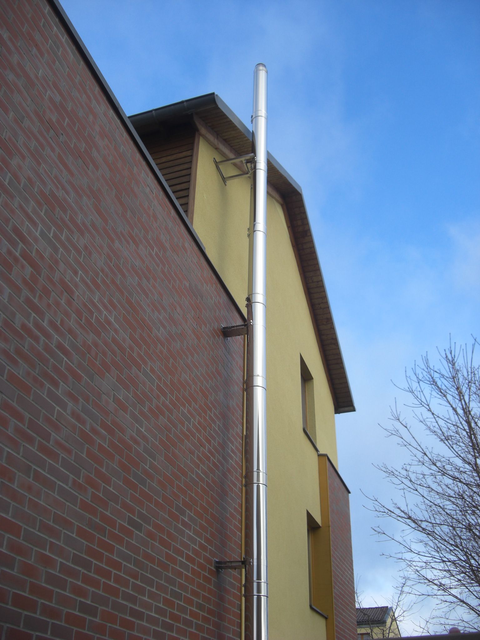Edelstahlschornstein an einer gelben Hauswand und einer Ziegelwand im Vordergrund