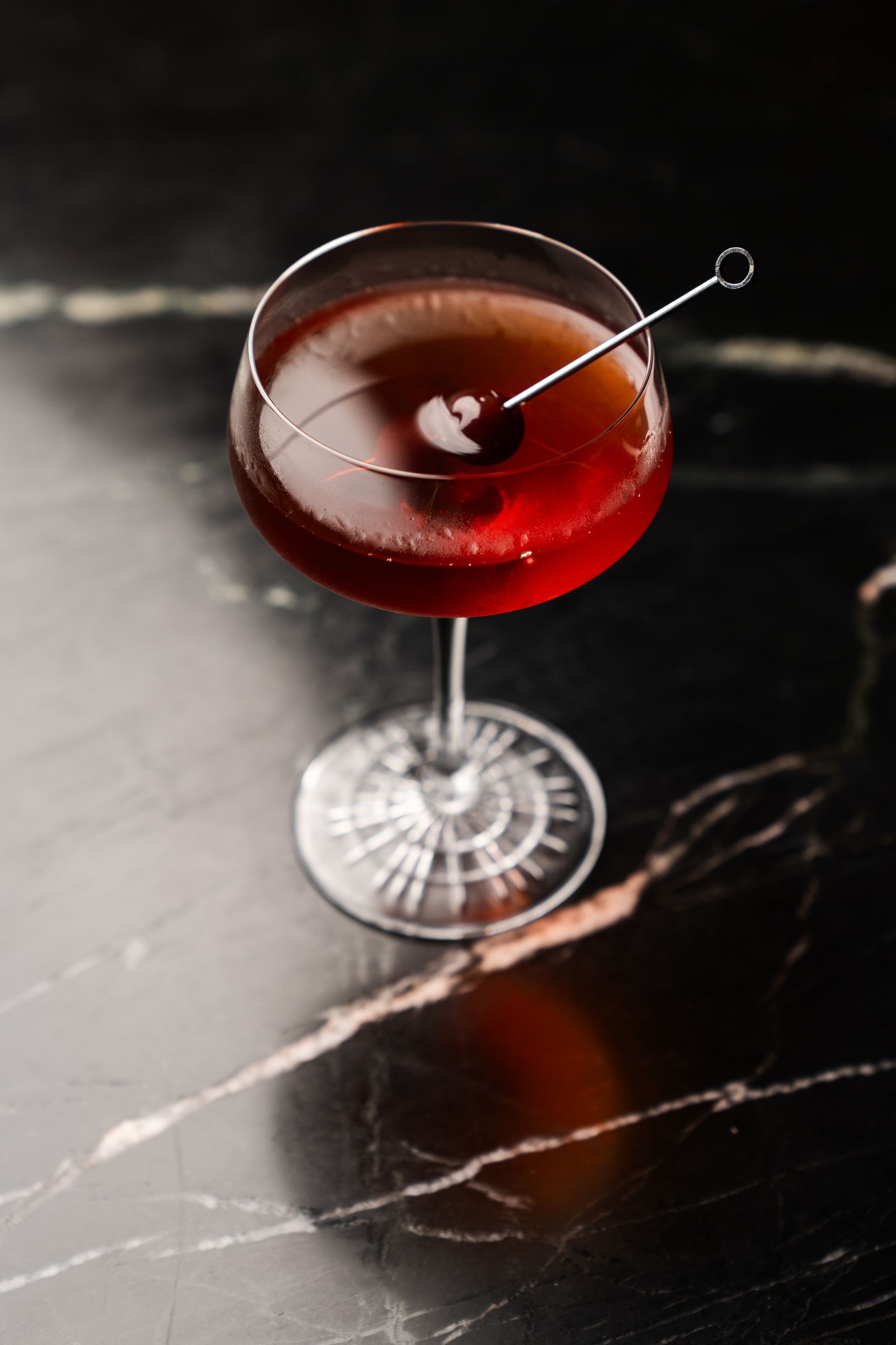 Eine Nahaufnahme eines Cocktails mit einer Kirsche auf einem silbernen Zahnstocher auf einem schwarzen Marmor Tisch.