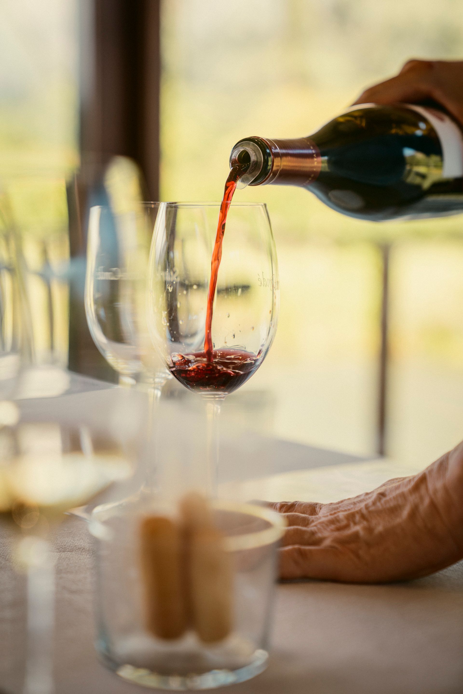 Tisch mit Weingläsern bei einer Weinprobe. In ein Glas wird Rotwein eingegossen.