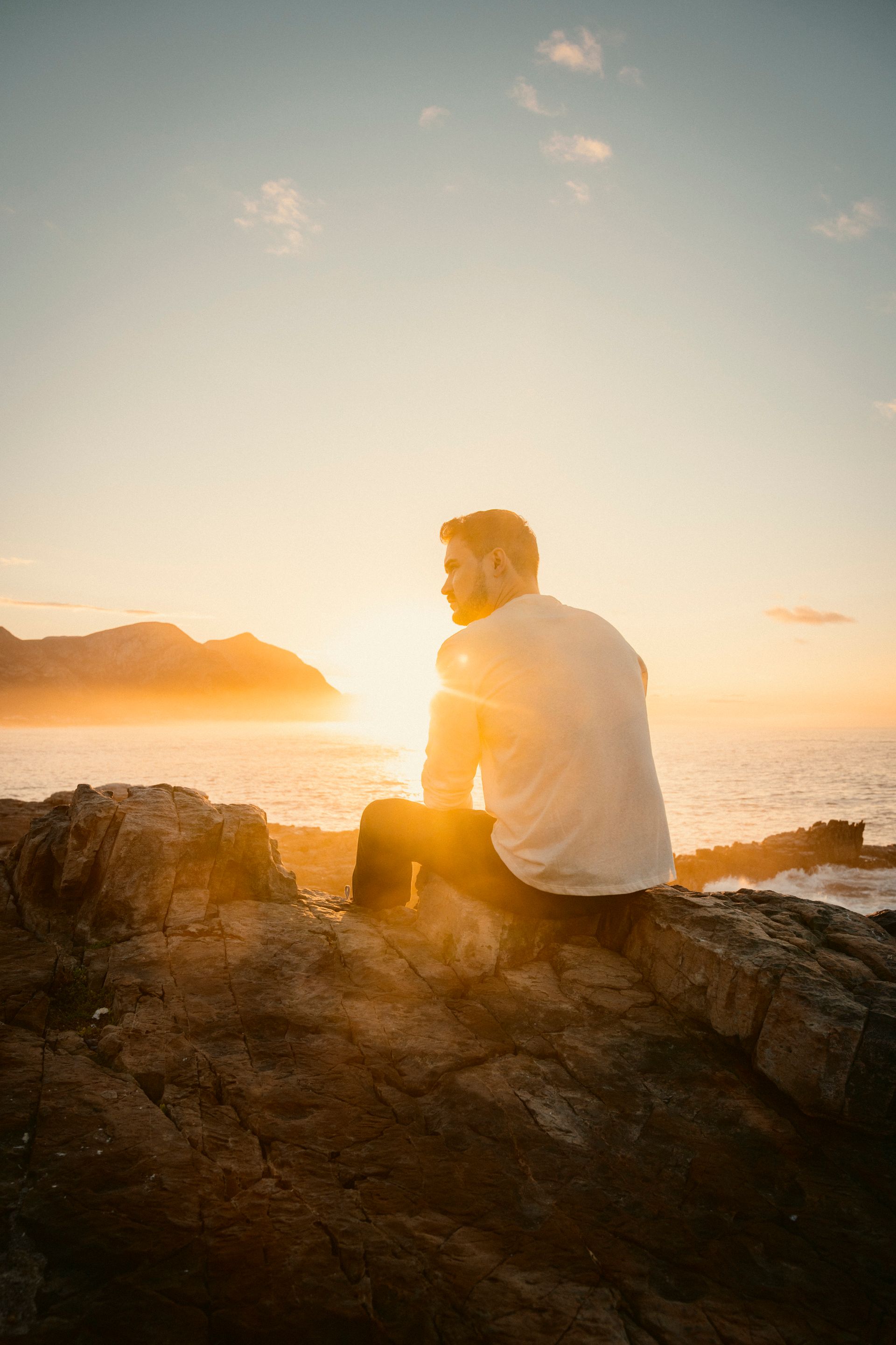 Markus sitzt im weißen T-Shirt auf einem Felsen und schaut über das Meer bei goldener Sonnenuntergangs Stimmung