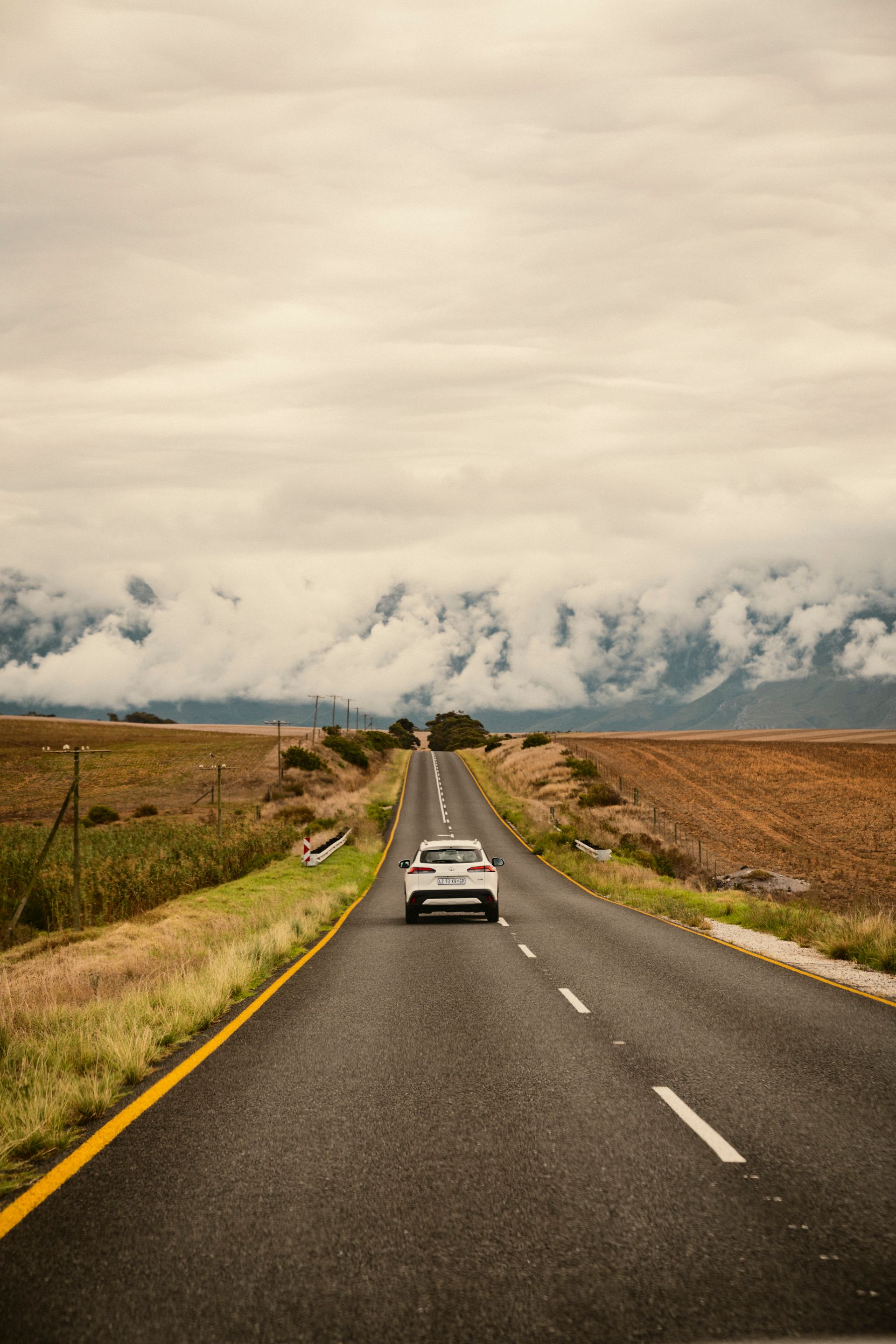 Bick über Landstraße auf ein weißes Auto. Rechts und links gelb orangene Flächen im Hintergrund wolkenverhangene Berge