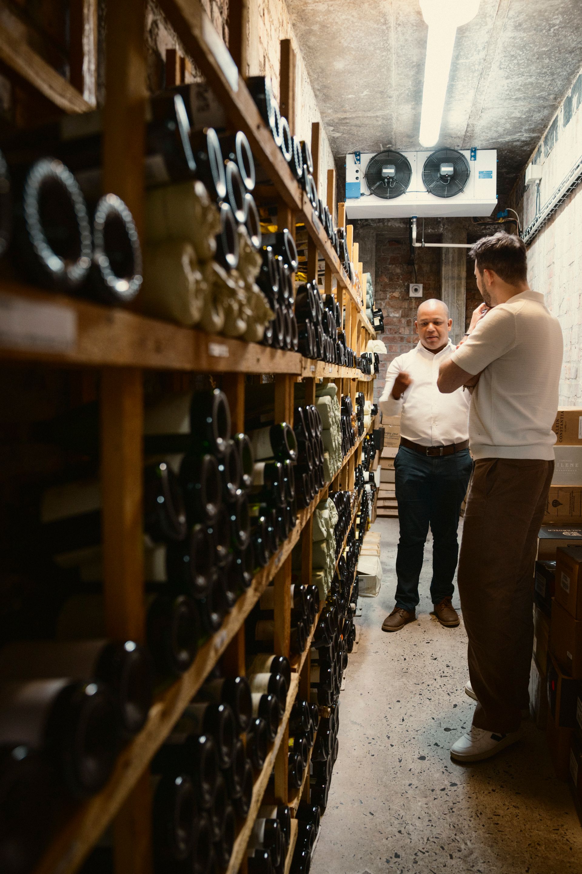 Markus und ein Mann unterhalten sich im Weinlager des Weingutes vor einem regal voll mit liegenden Weinflaschen