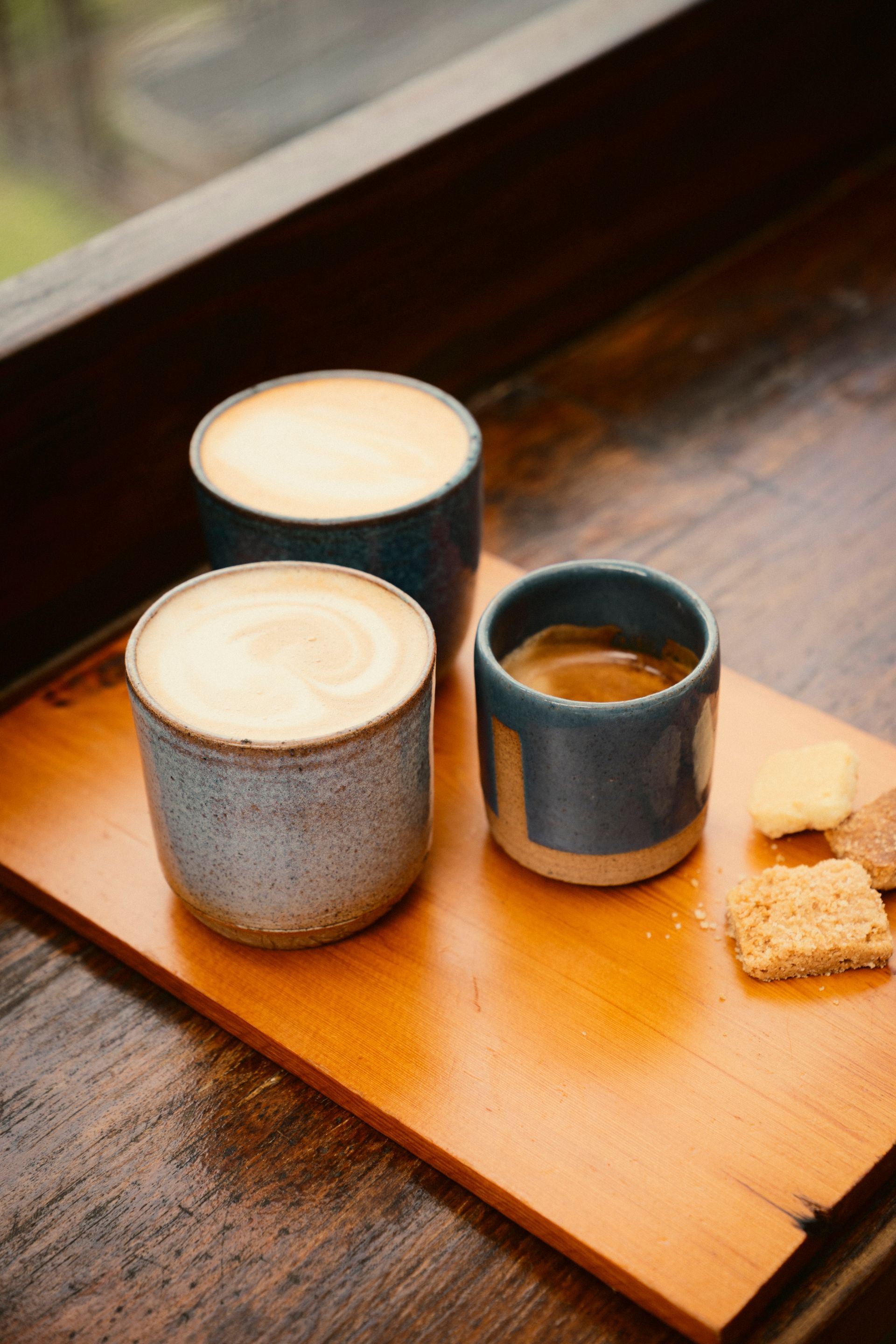 Holzbrett mit drei Henkellosen Kaffeebechern. Zwei mit Milchschaum einer mit Crema. Daneben liegen kleine Kekse
