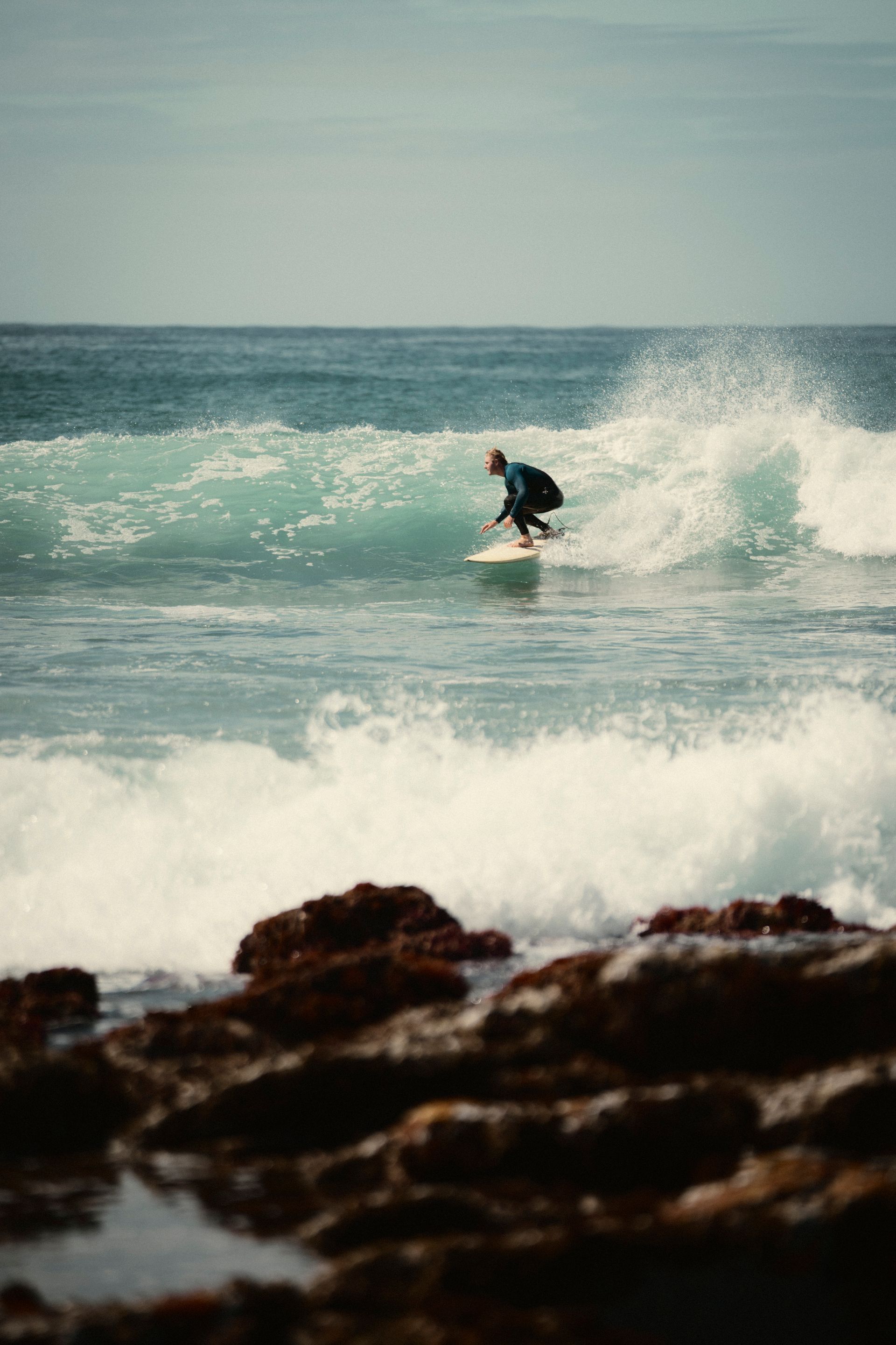 Blick auf das Meer. Ein Surfer reitet eine Welle, im Vordergrund bricht noch eine Welle