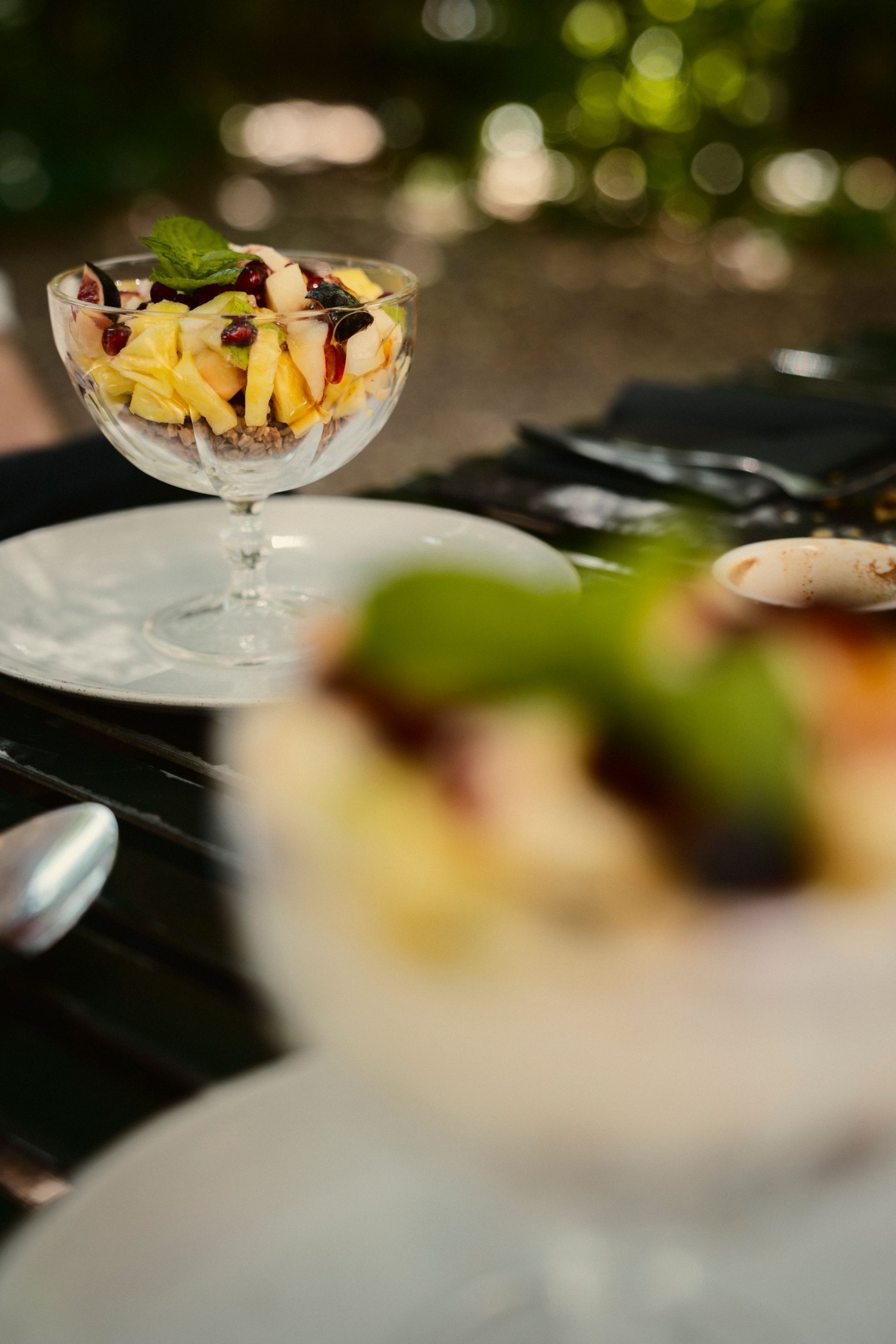 Zwei Hohe Glasschalen auf weißen Tellern mit Obstsalat und Minzblatt. Die schale im Vordergrund ist unscharf