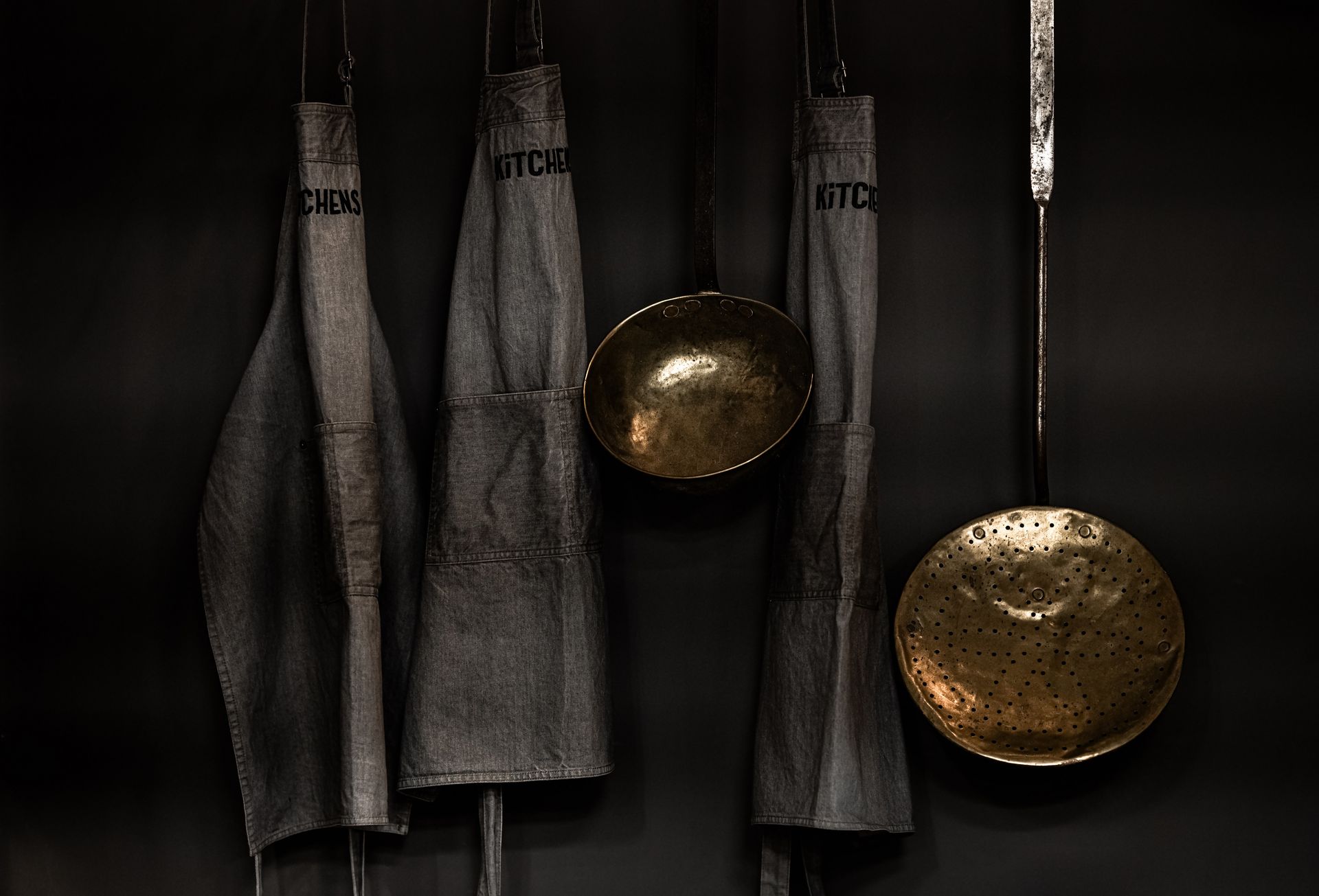 Graue KITCHENS Schürzen hängen an einer dunklen Wand neben goldfarbenen großen Schöpfkellen