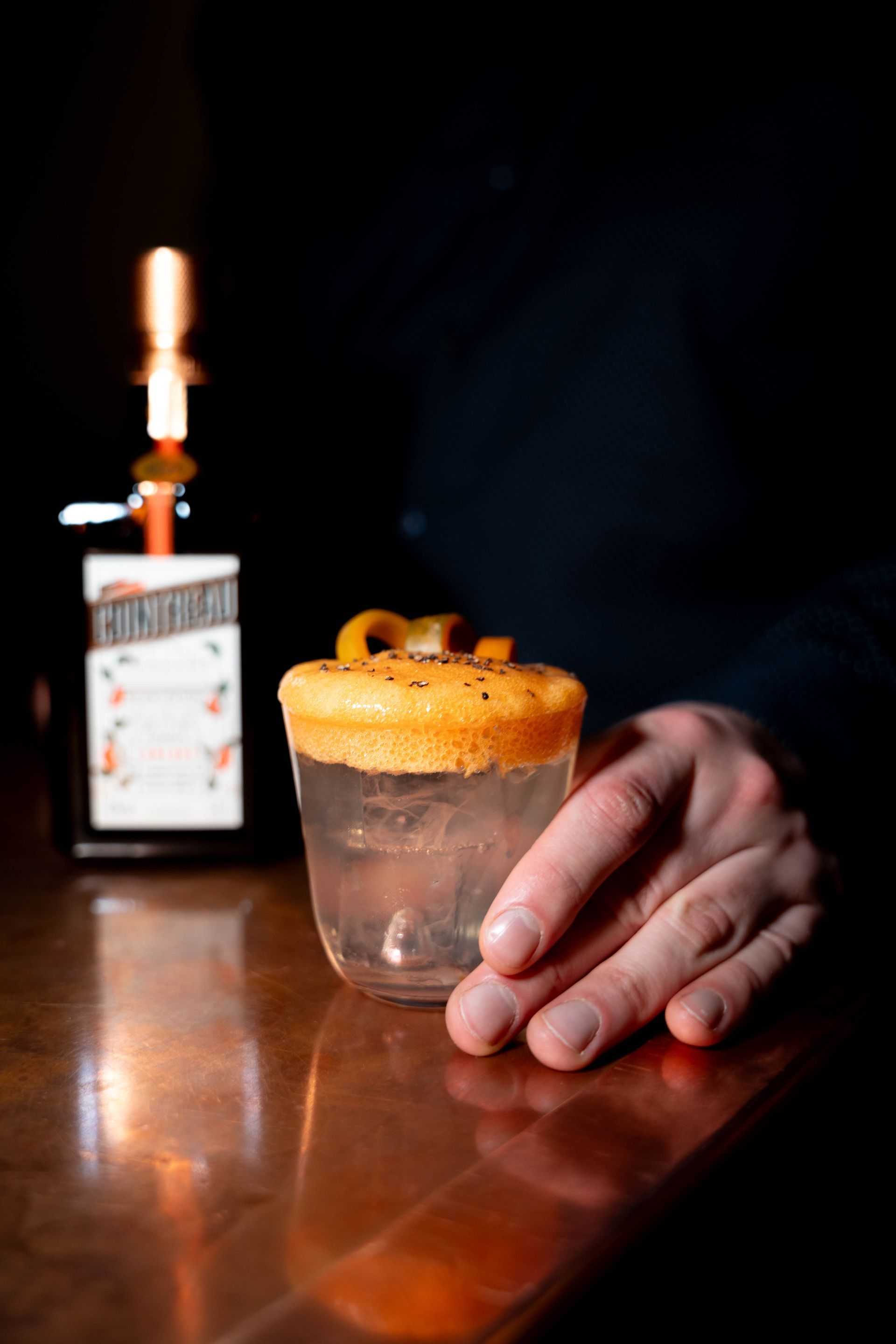 Eine Person hält einen Cocktail mit einer Orangenschale darauf. Im Hintergrund steht eine Cointreau Flasche