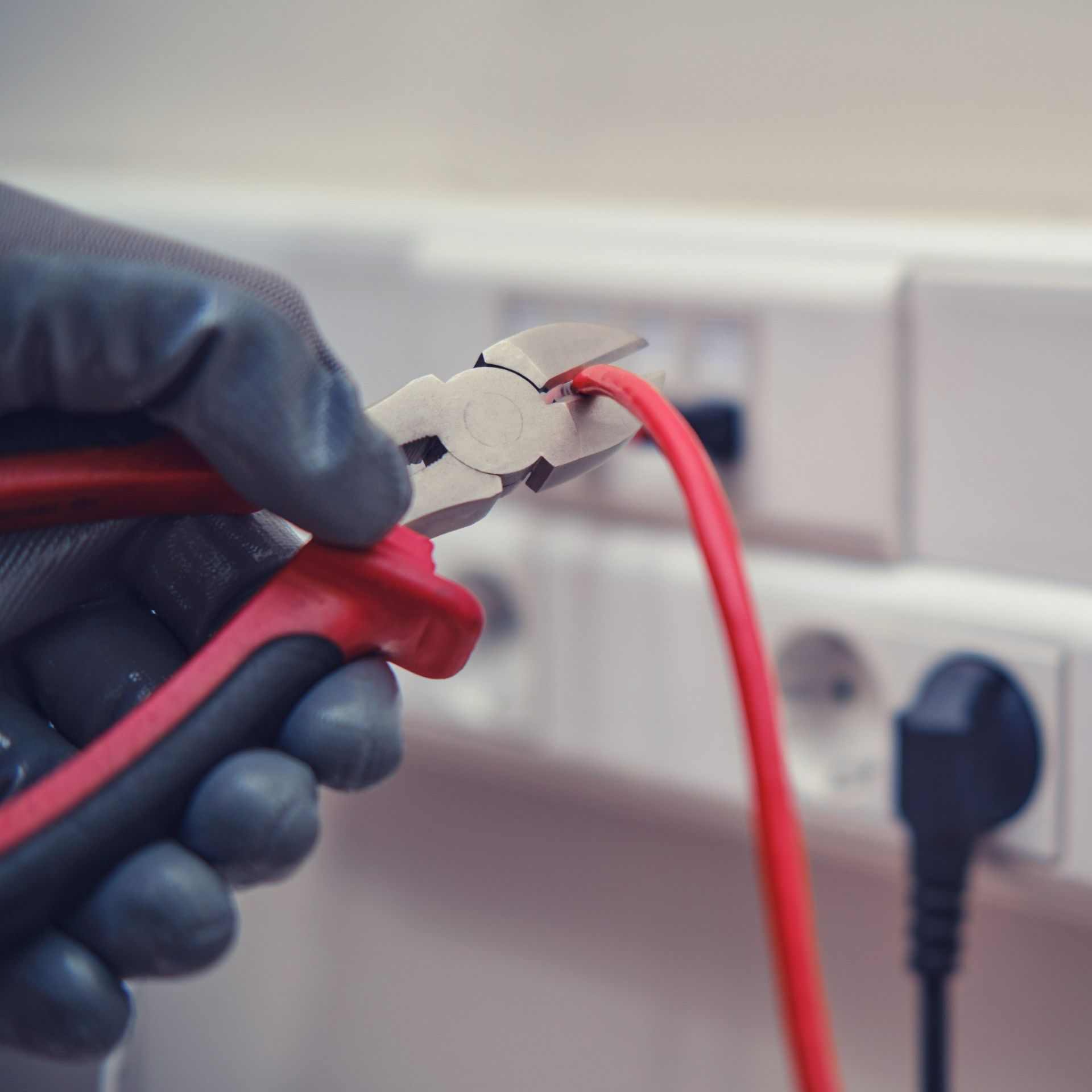 Mano enguantada que utiliza alicates rojos para cortar un cable eléctrico rojo cerca de los enchufes de pared.