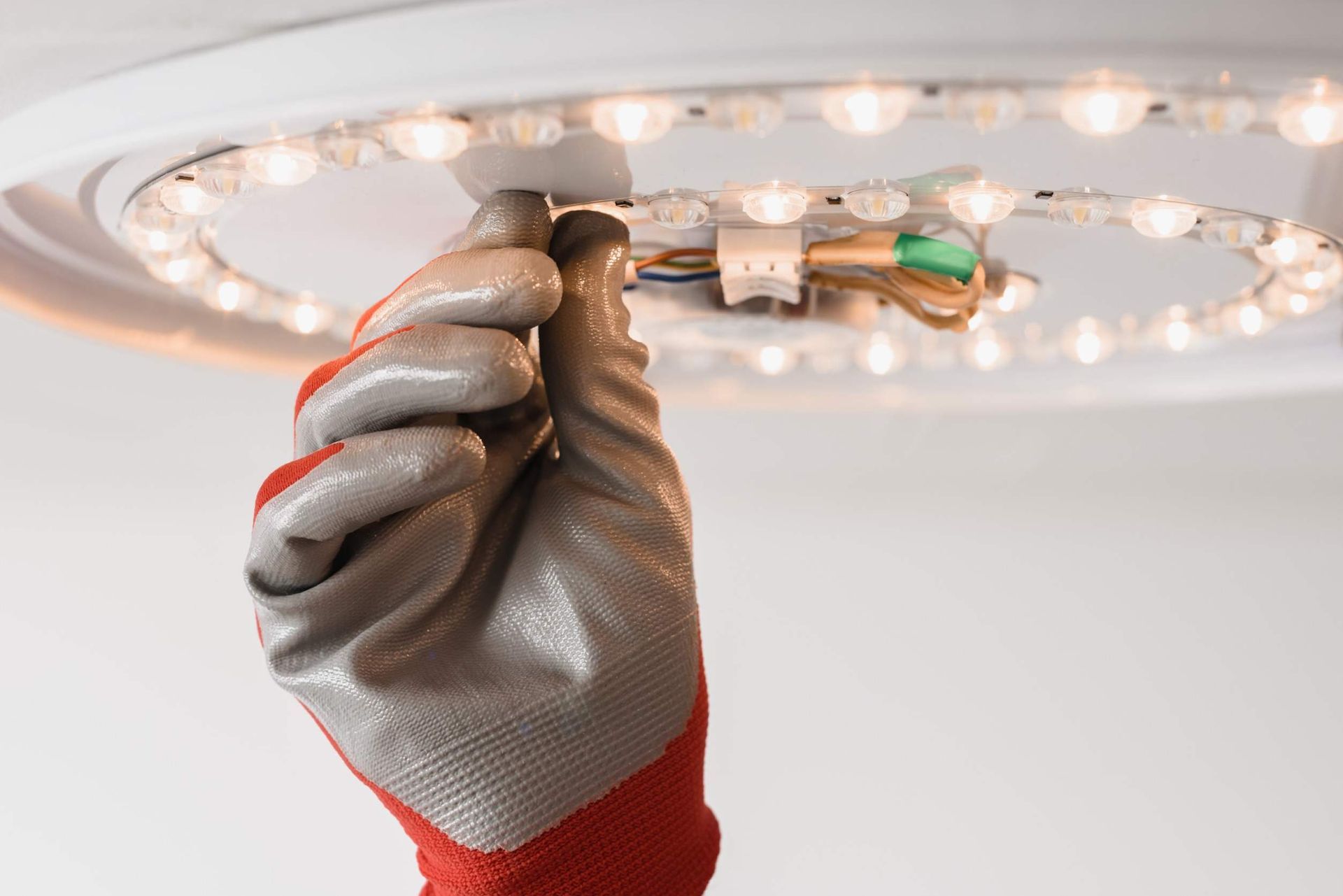 Mano enguantada conectando cables a una lámpara LED circular. Techo blanco, atención al trabajo eléctrico.