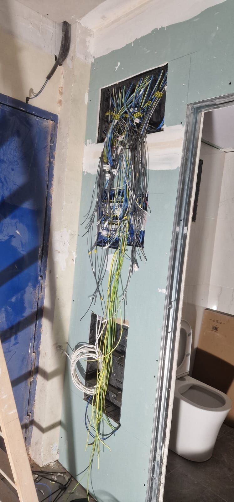 Un panel eléctrico en construcción, con cables expuestos que sobresalen de la pared. La pared es azul claro y se ve una puerta azul a la izquierda.