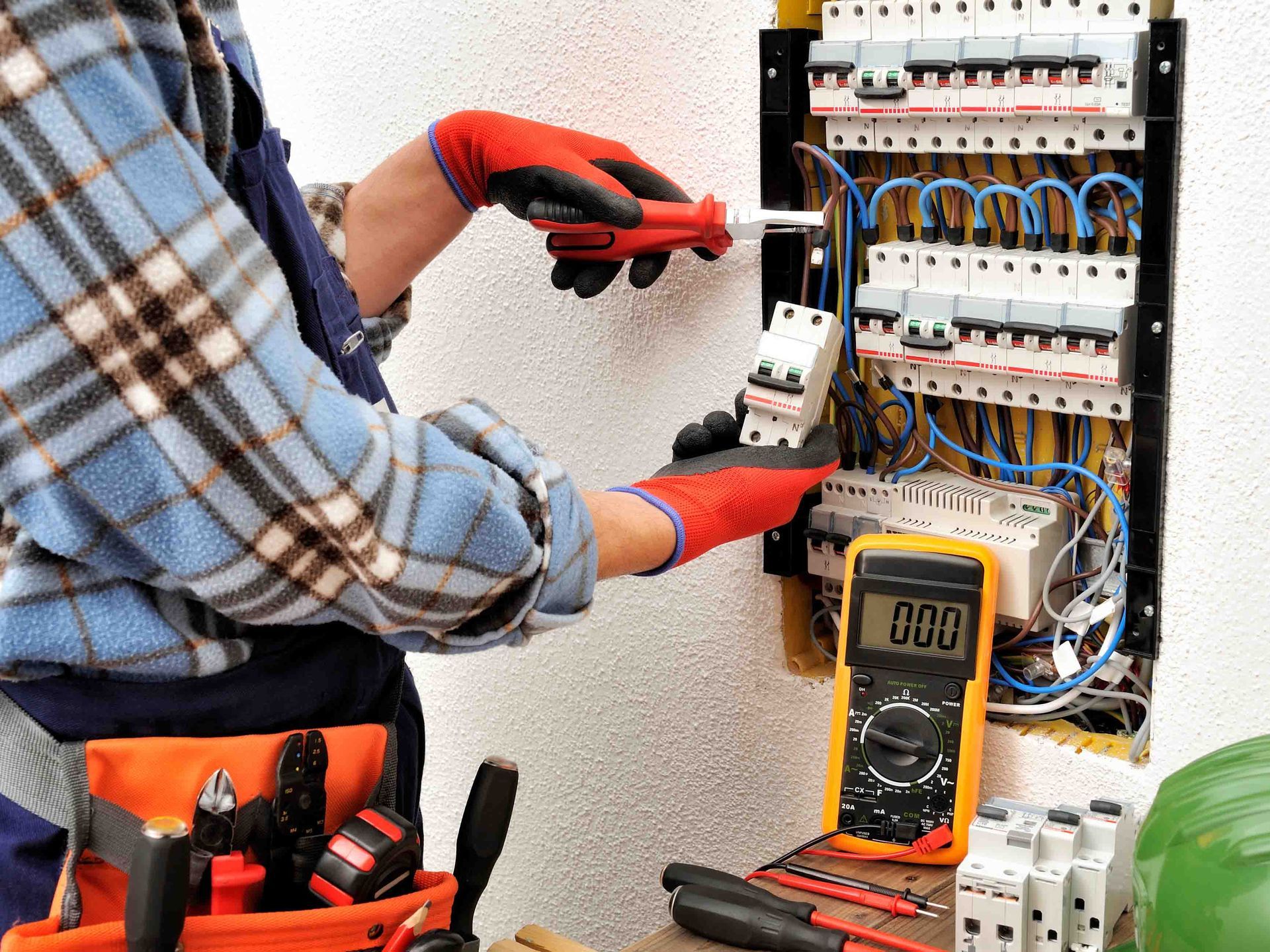 Electricista con camisa a cuadros azul instalando componentes eléctricos en un panel, usando guantes rojos y usando un destornillador.