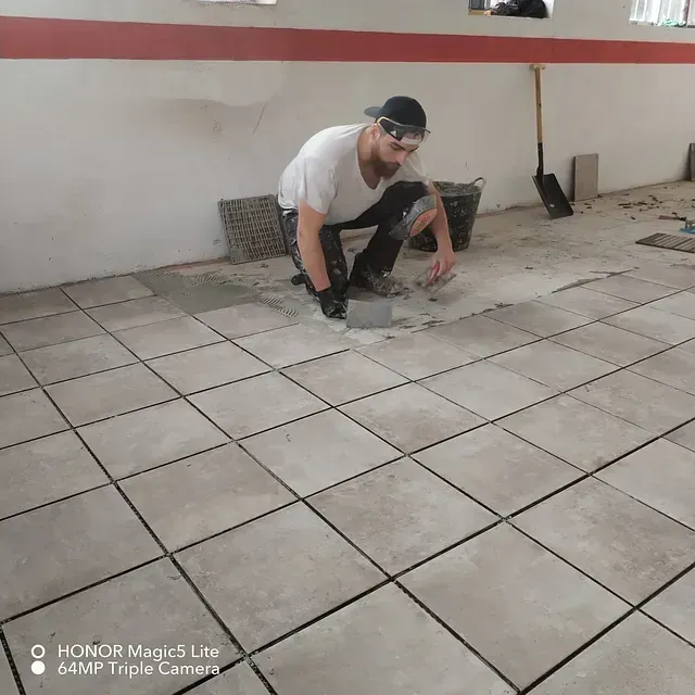 Un hombre está arrodillado sobre un suelo de baldosas usando una pala.