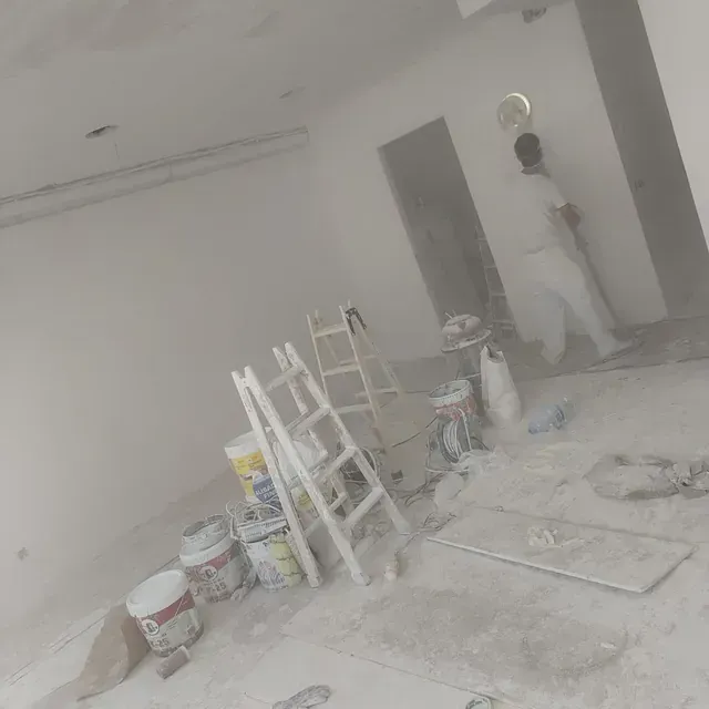 Un hombre está de pie en una habitación con baldes de pintura y una escalera.