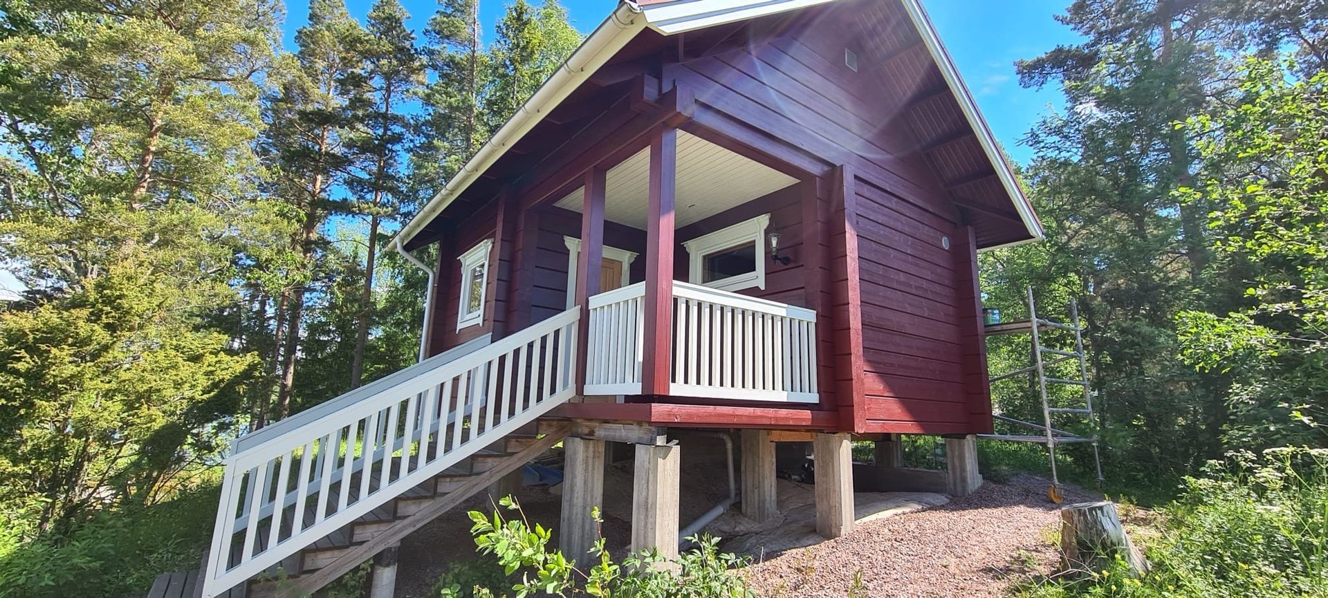 Pieni punainen mökki paalujen päällä metsäisellä alueella, jossa on valkoinen portaikko ja kuistin kaide.