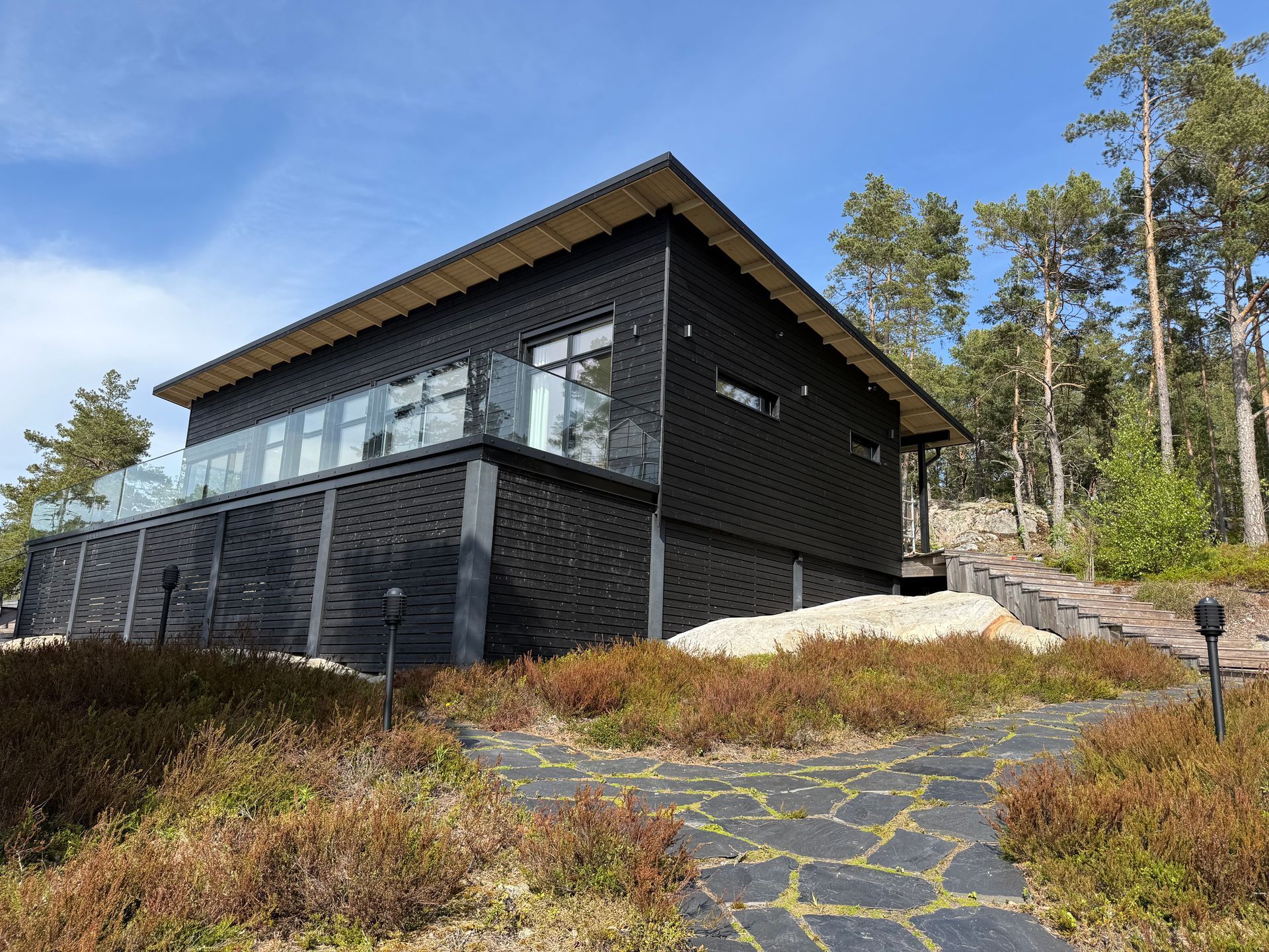 Moderni musta talo suurilla ikkunoilla metsäisellä rinteellä, katsottuna kivipolulta sinisen taivaan alla