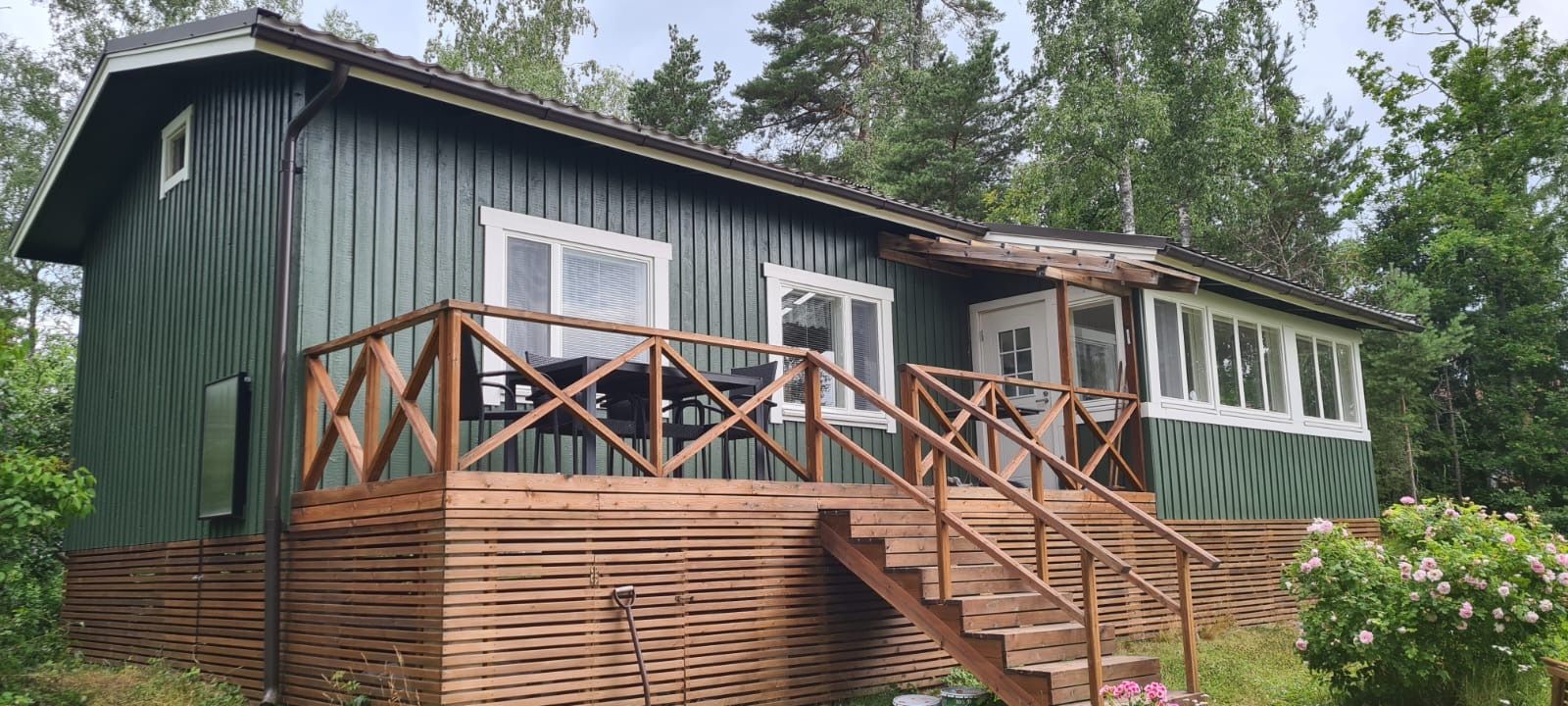 Vihreä mökki, jossa valkoiset ikkunat, puiset kuistiportaat ja ristikkohelma metsäisessä ympäristössä