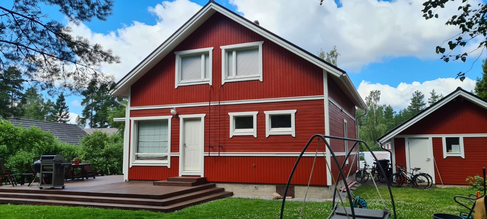 Punainen puutalo valkoisilla reunuksilla ja keinulla vehreällä pihalla sinisen taivaan alla