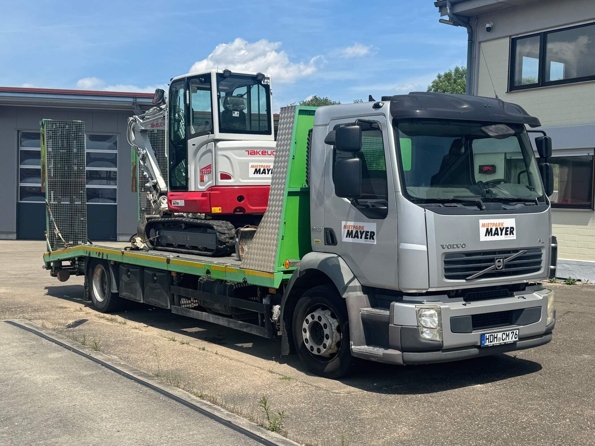 Ein grauer Lkw mit einer grünen Ladefläche, auf der ein kleiner rot-weißer Kettenbagger geladen ist