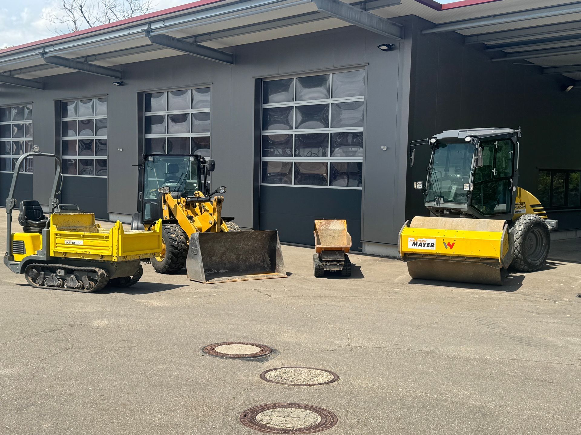 Kleine gelbe Baumaschinen wie Dumper, Radlader und Walze aufgereiht vor einer Halle.