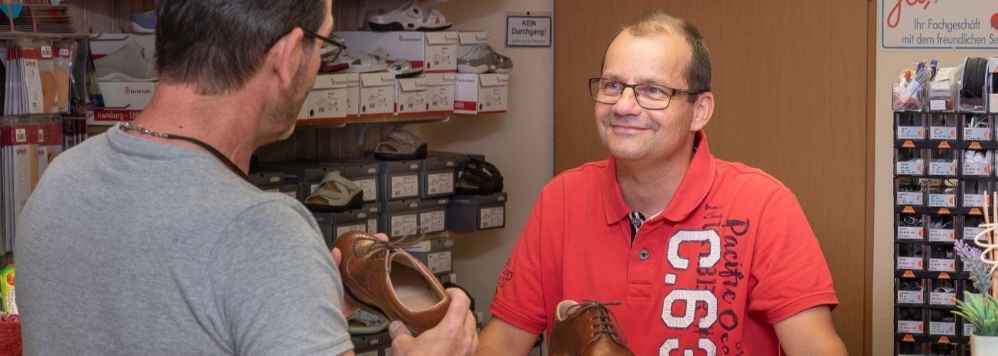 Ein Mann in einem roten Hemd spricht mit einem anderen Mann, der ein Paar Schuhe hält.