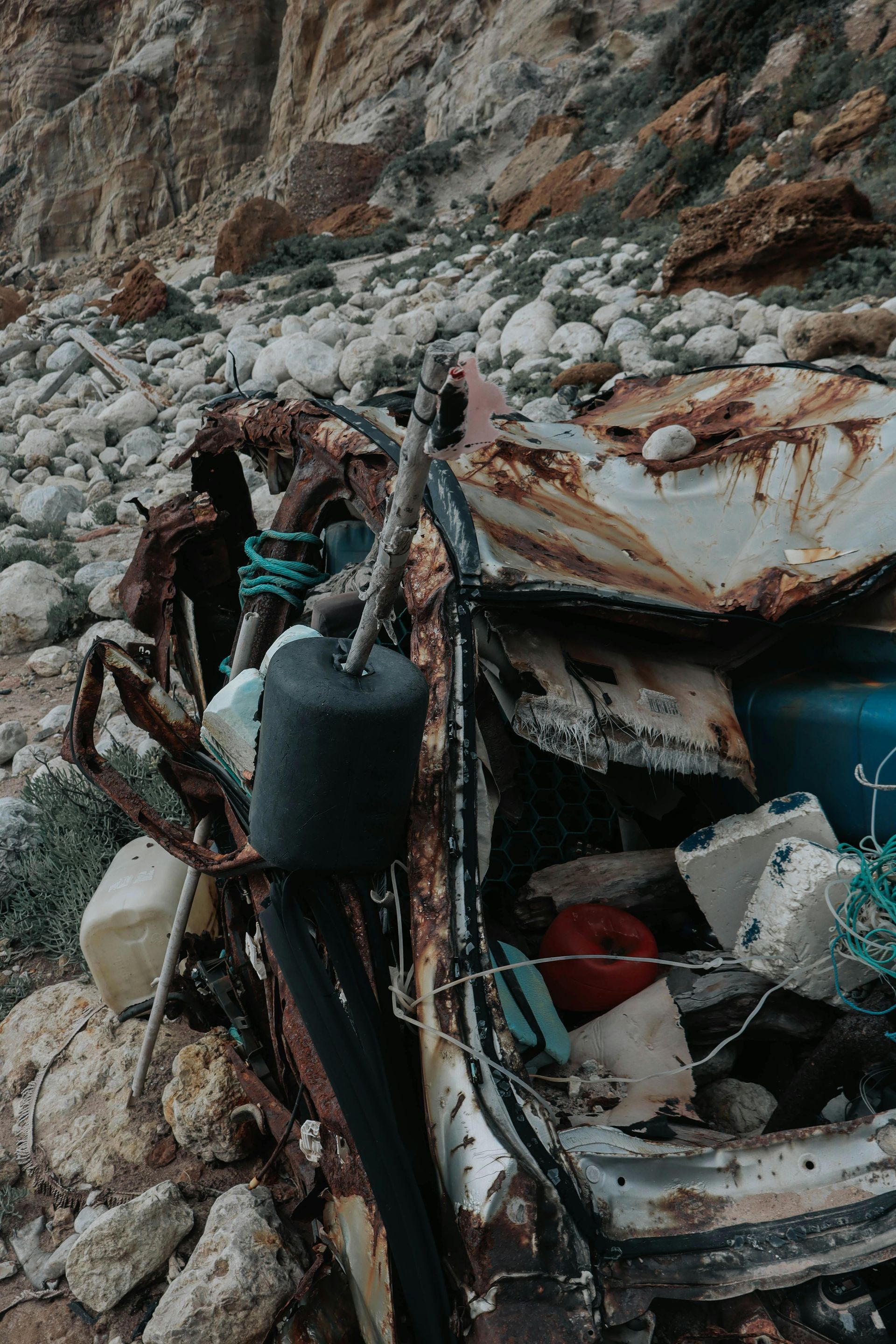 Embarcación dañada entre escombros rocosos en la costa, con un contenedor azul y basura esparcida.