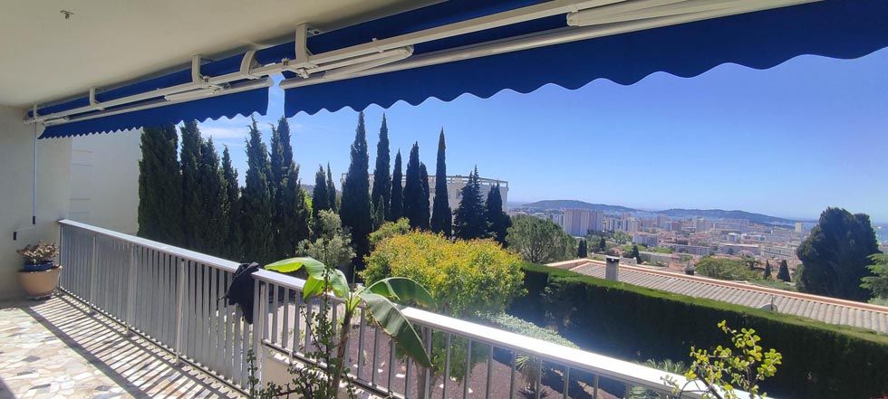 Balcon avec un auvent bleu, avec vue sur une ville et des arbres par une journée ensoleillée.