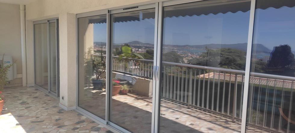 Portes coulissantes en verre donnant sur un paysage urbain avec un balcon, une balustrade et un ciel ensoleillé.