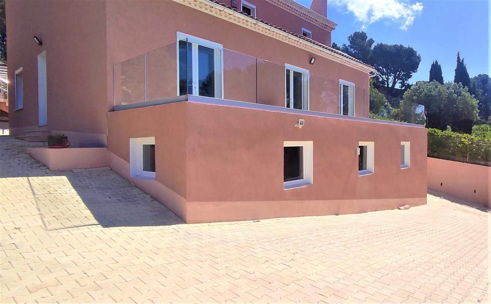Maison de deux étages de couleur saumon avec un balcon en verre et plusieurs fenêtres à cadre blanc, située sur une allée en pierre.
