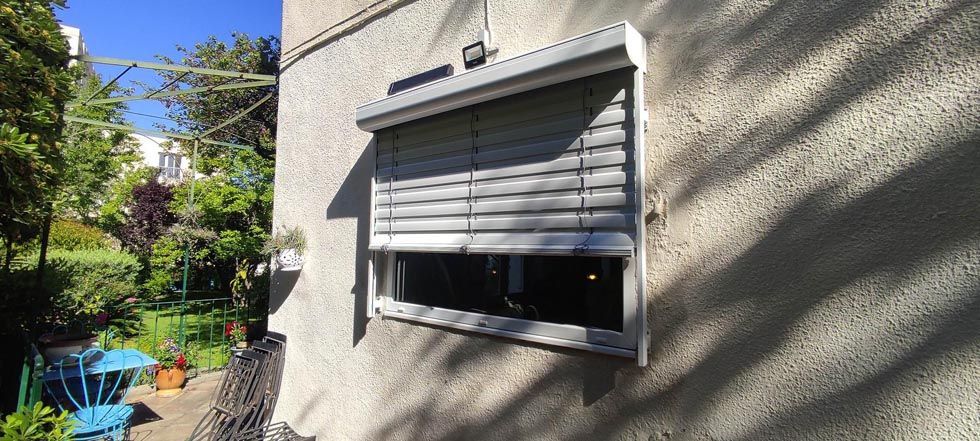 Fenêtre avec stores partiellement fermés sur un mur en stuc. Un jardin est visible en arrière-plan.