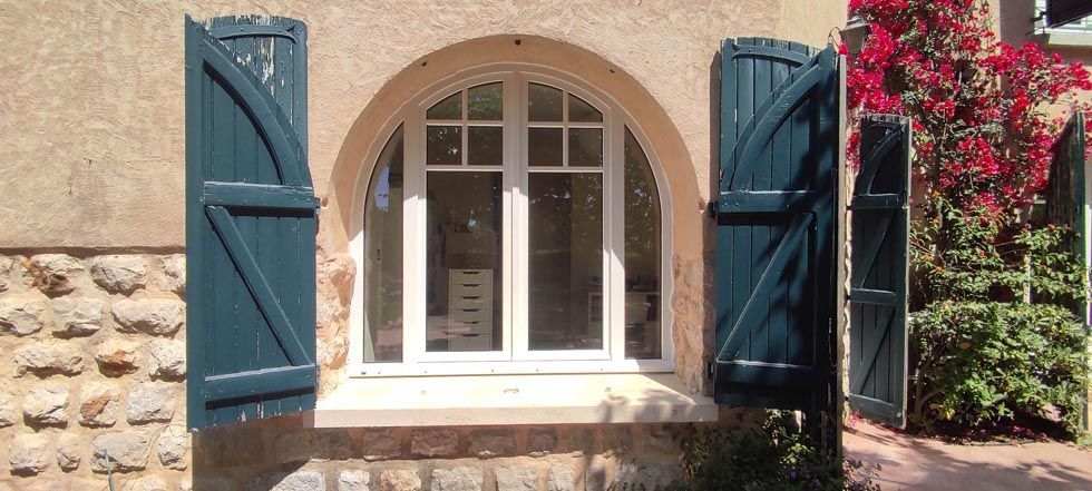 Fenêtre cintrée aux volets bleus sur un mur de pierre, entourée de plantes fleuries.