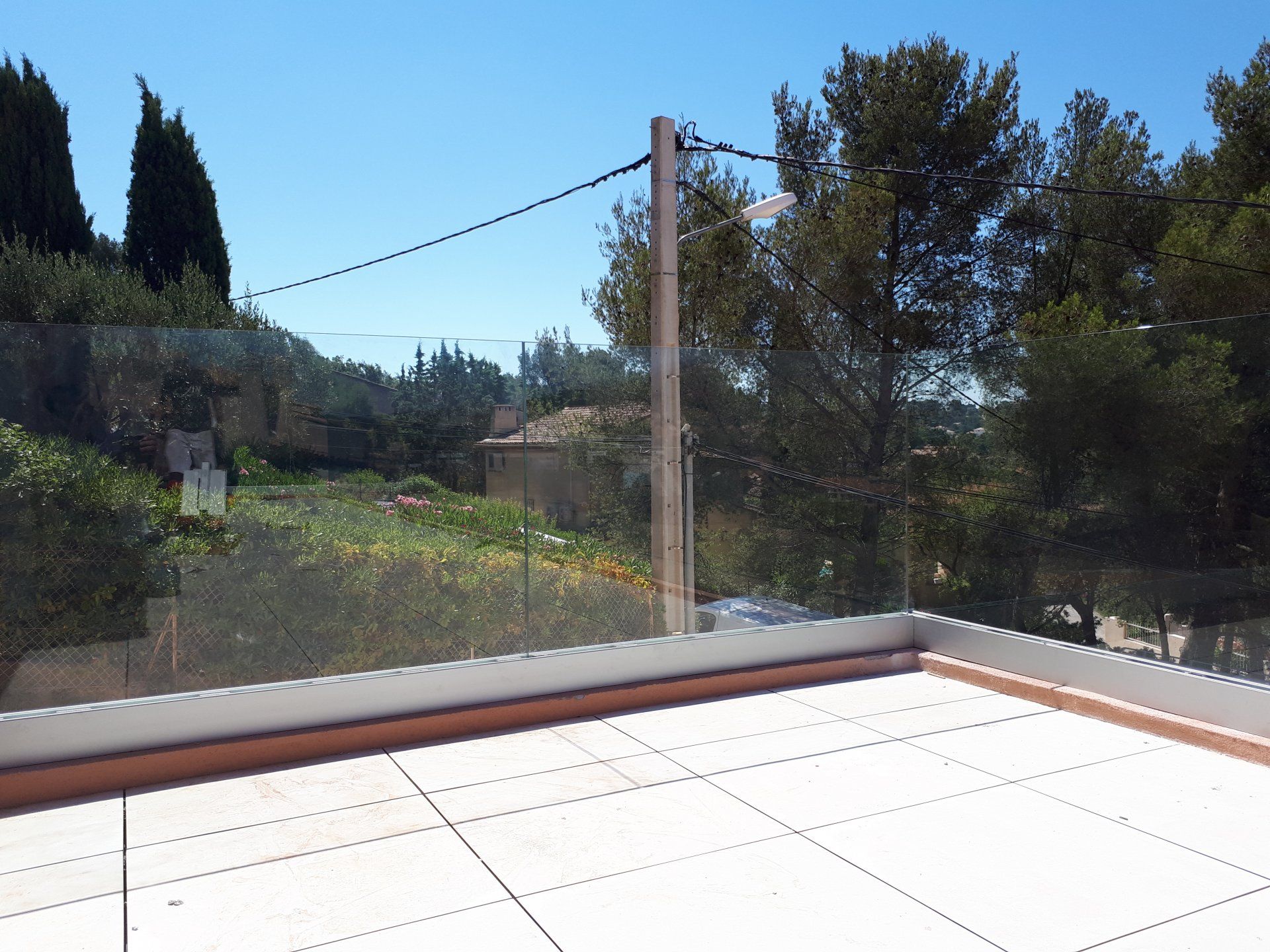 Terrasse avec balustrade en verre donnant sur des arbres, un bâtiment lointain et un ciel bleu vif.