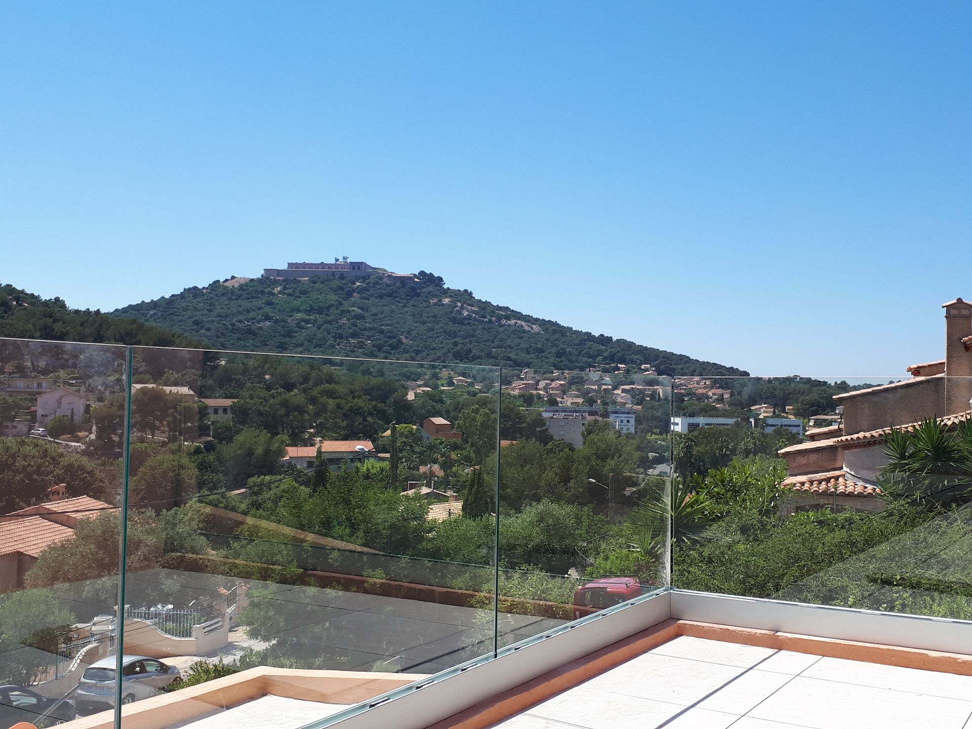 Vue d'une ville à flanc de colline sous un ciel bleu, avec ses toits en terre cuite et sa végétation luxuriante. Un balcon vitré se trouve au premier plan.