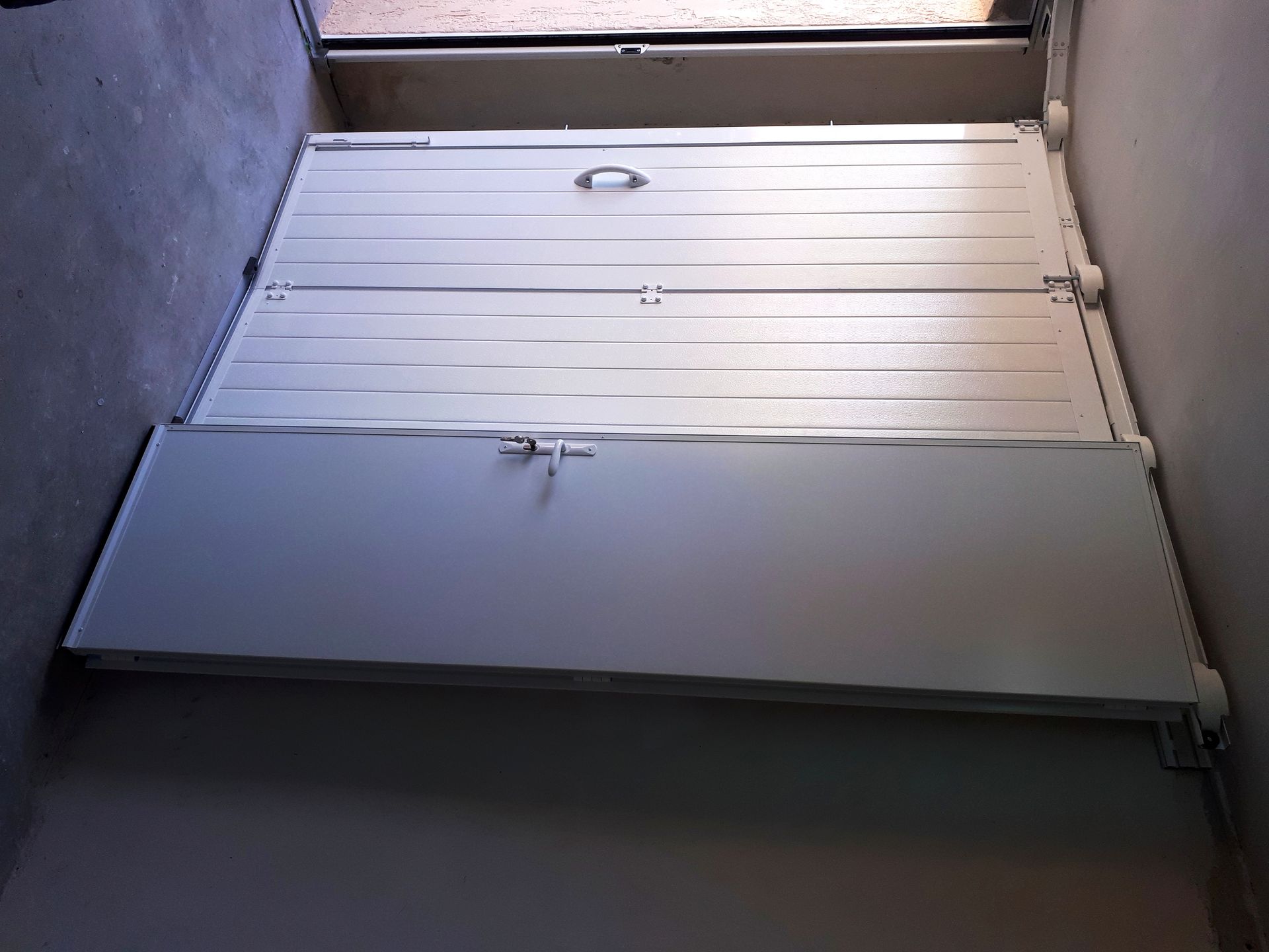 Porte pliante en métal blanche, avec poignée et loquet, fermée. Installée dans une porte étroite.