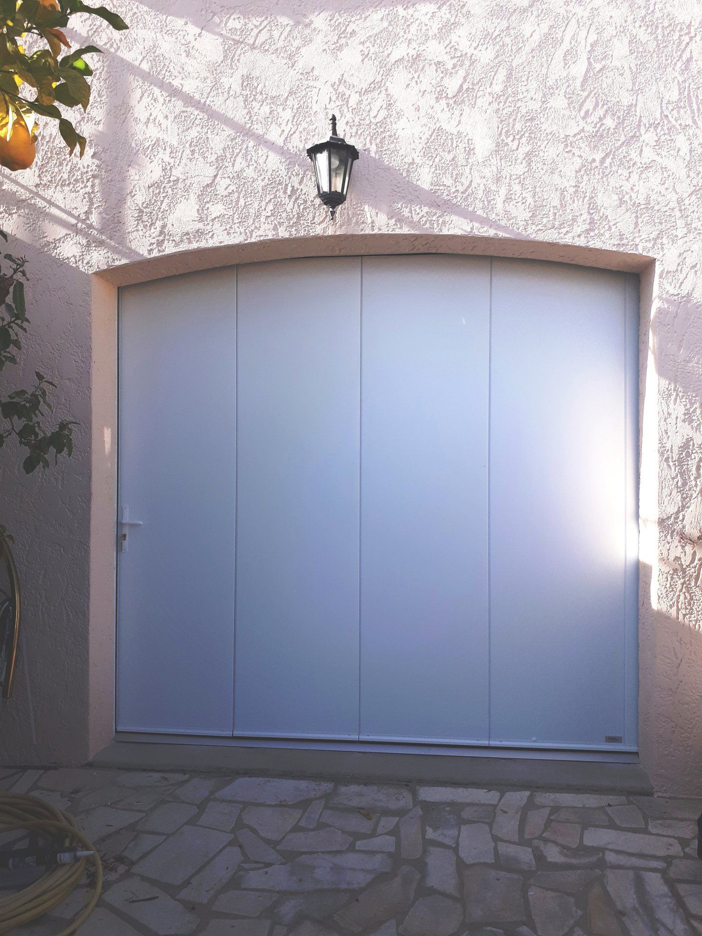 Porte de garage fermée de couleur claire, avec panneaux verticaux encastrés dans un mur en stuc. Ci-dessus, une lanterne noire.