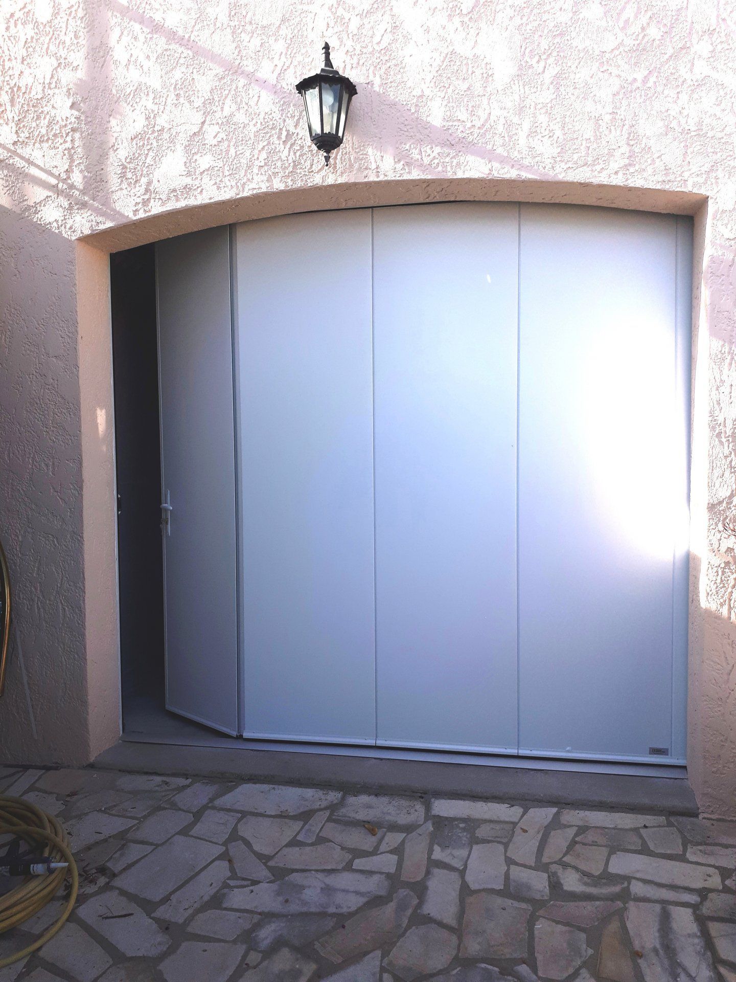Porte de garage gris clair à plusieurs panneaux, partiellement ouverte. Installée sur une terrasse en pierre texturée.