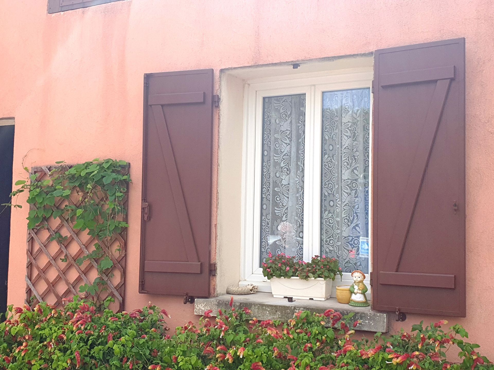 Fenêtre aux volets marron sur un mur couleur saumon, avec un treillis et des fleurs en dessous.