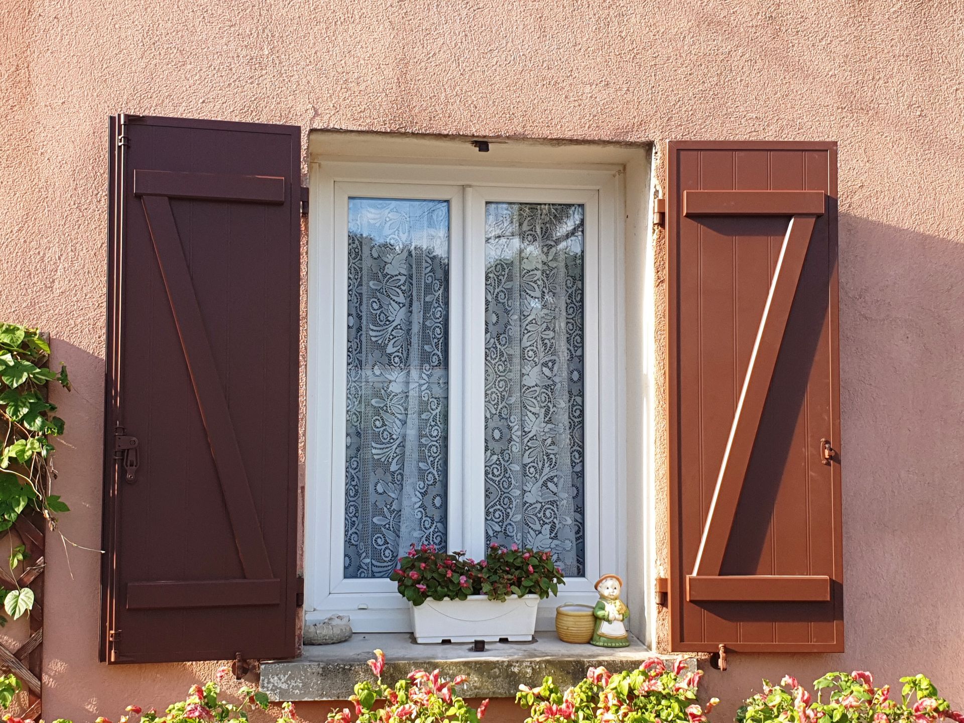 Fenêtre avec volets marron, cadre blanc, rideaux de dentelle et jardinière sur un mur en stuc rose.