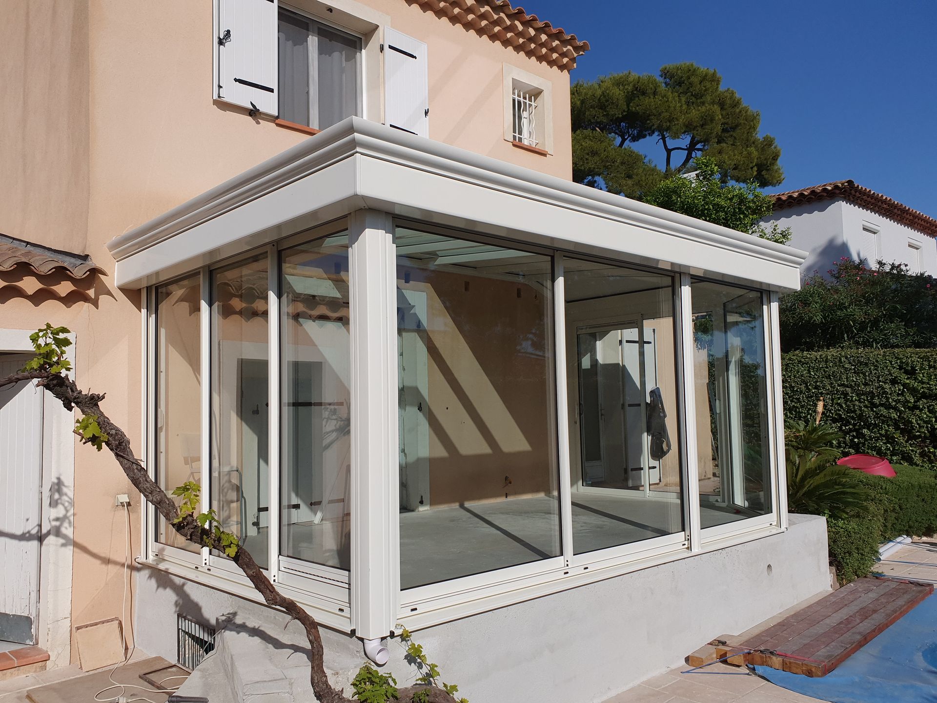 Véranda à cadre blanc, attenante à une maison beige, avec fenêtres en verre. Située à l'extérieur, elle bénéficie d'un ensoleillement intense.