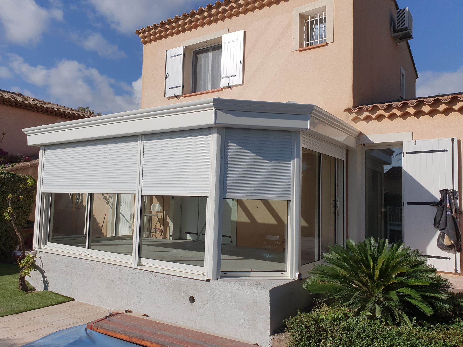 Véranda blanche attenante à une maison beige, avec stores fermés et verdure environnante.