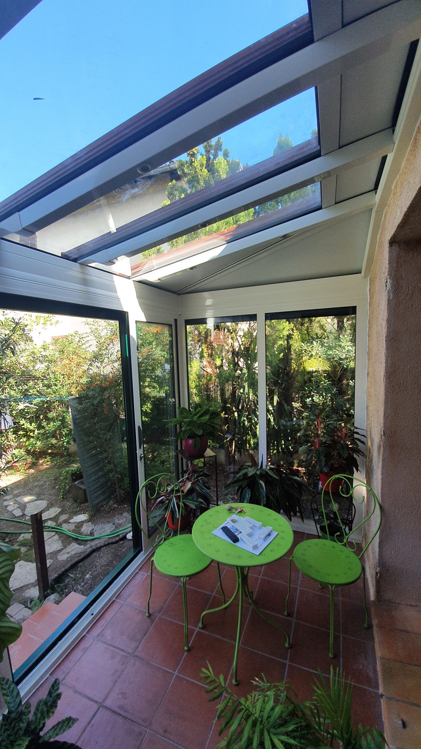Véranda avec plafond en verre, table verte et tabourets, entourée de plantes et vue sur le jardin.