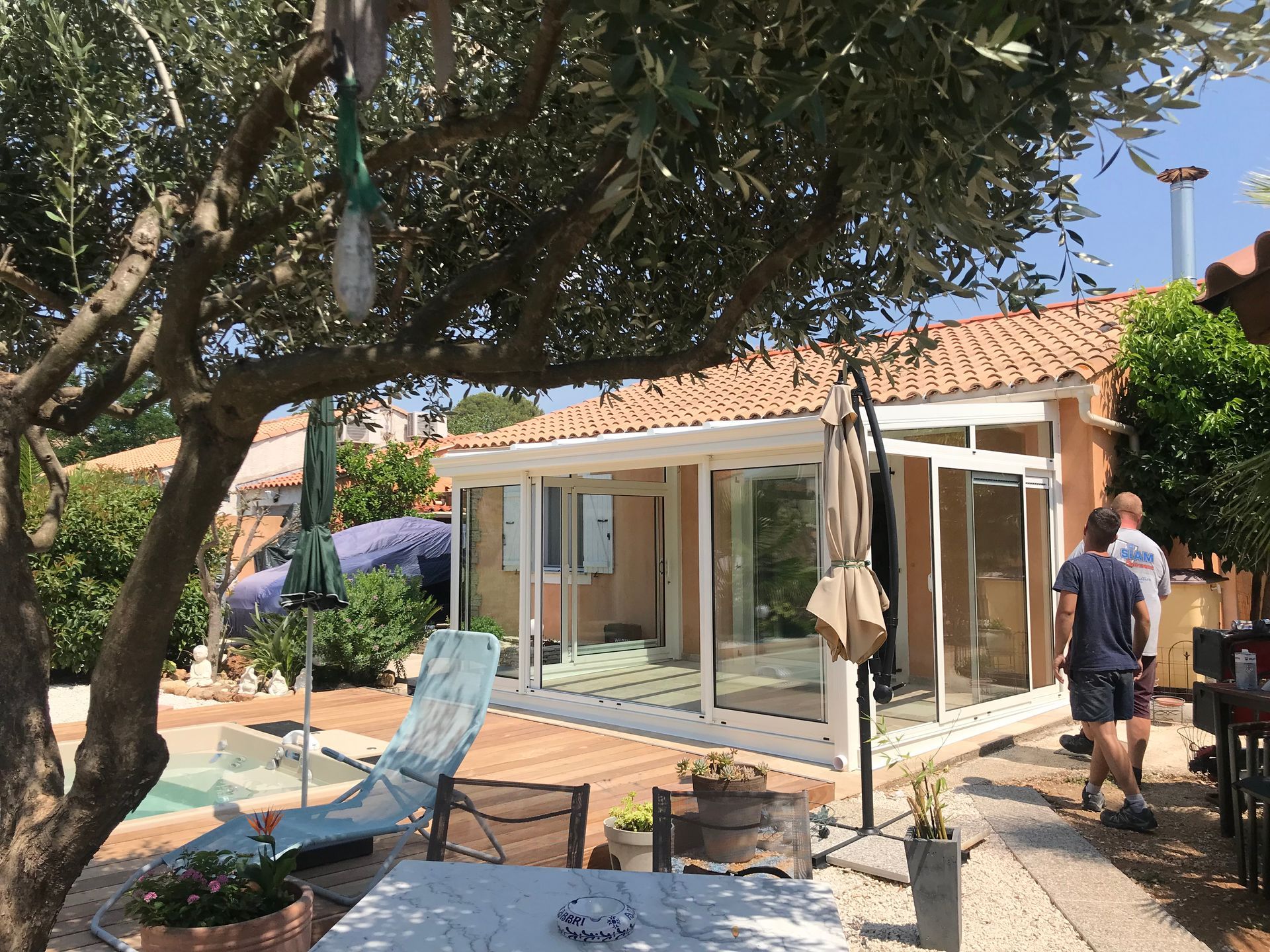 Scène extérieure ensoleillée avec une structure aux parois de verre, une piscine et deux personnes à proximité.