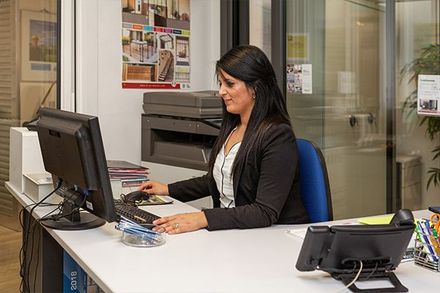 Une femme en blazer noir, assise à un bureau, qui tape sur un ordinateur