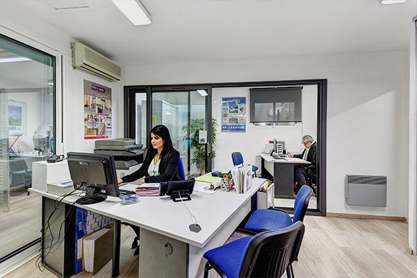 Une femme à son bureau travaille sur ordinateur. Une autre personne à l'arrière-plan est assise à son bureau. Murs blancs, chaises bleues.