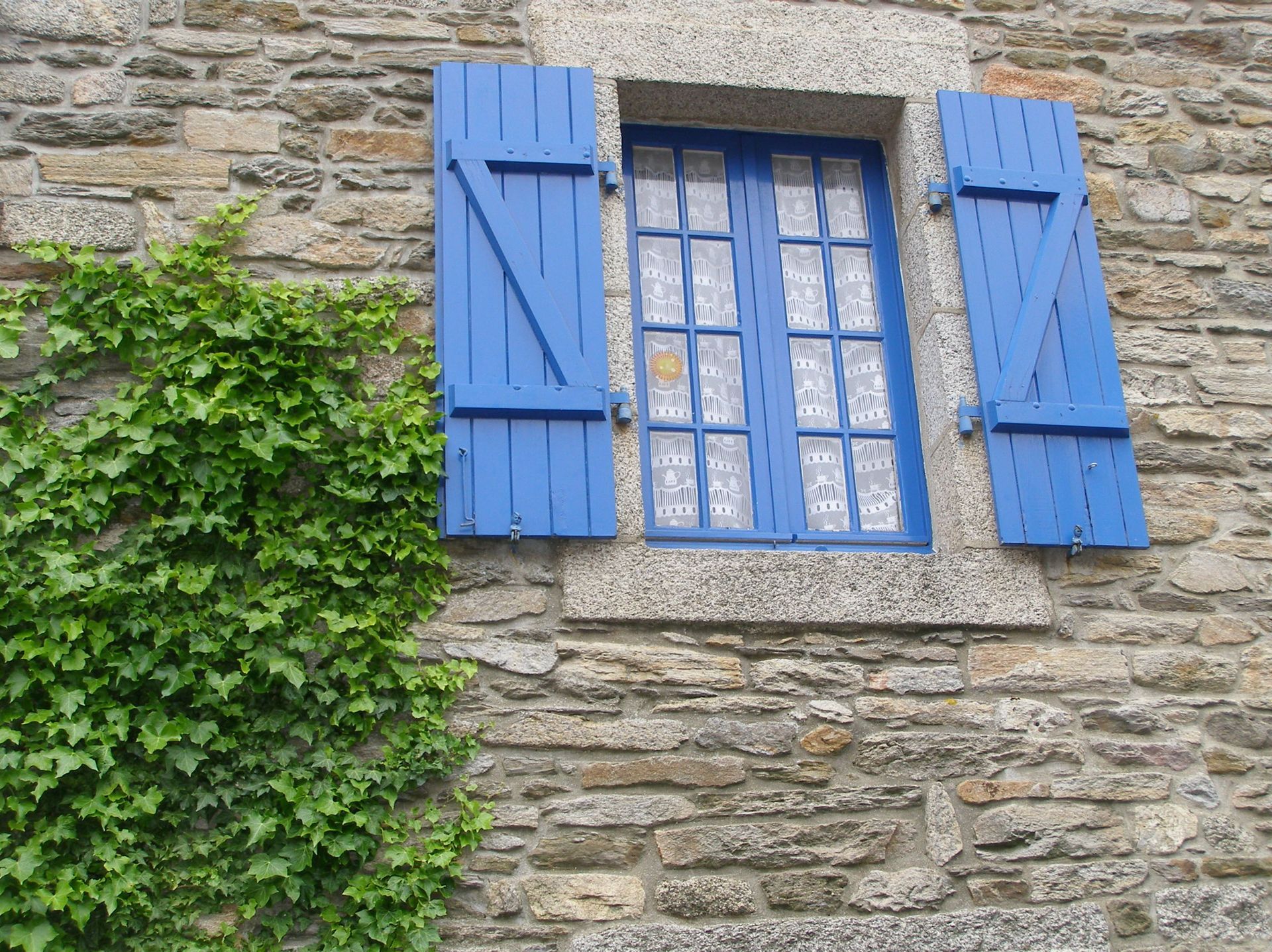 Un mur de pierre avec une fenêtre aux volets bleu vif, une vigne verte et feuillue pousse sur le côté gauche