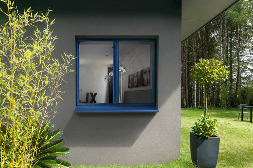 Fenêtre à cadre bleu sur un mur extérieur gris, donnant sur un jardin avec des arbres et des plantes en pot.