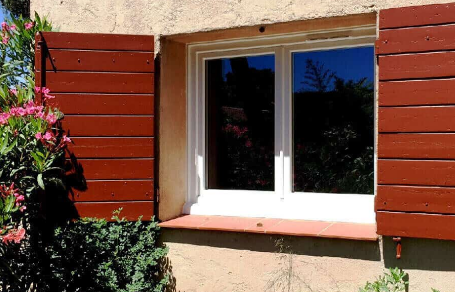 Fenêtre avec volets rouges et garnitures blanches, insérée dans un mur beige avec de la végétation sur les côtés.